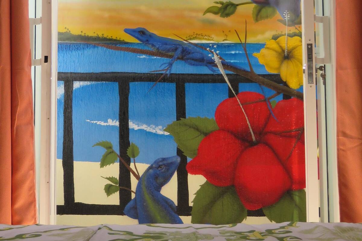 A room with a bed, a window with a view of palm trees, a blue lizard, and red flowers.