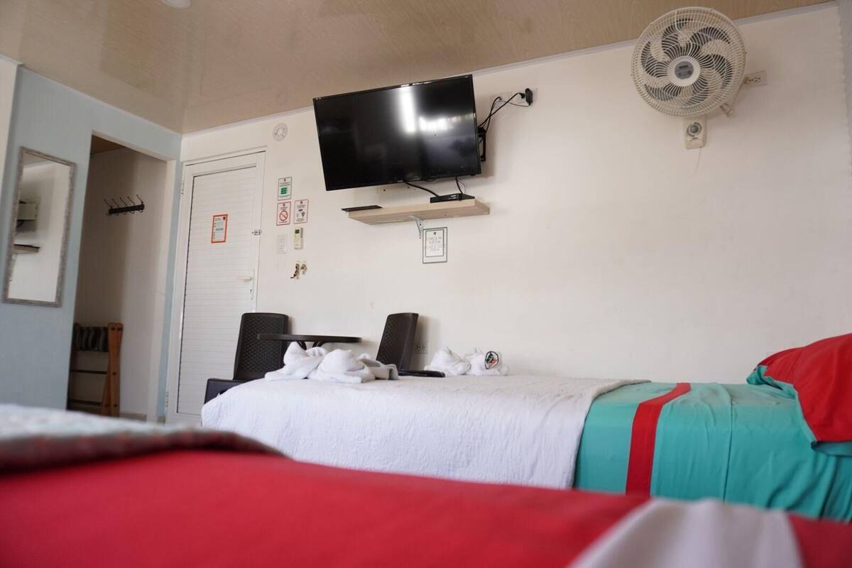A hotel room with a bed, a television mounted on the wall, a standing fan, and a desk with a chair.