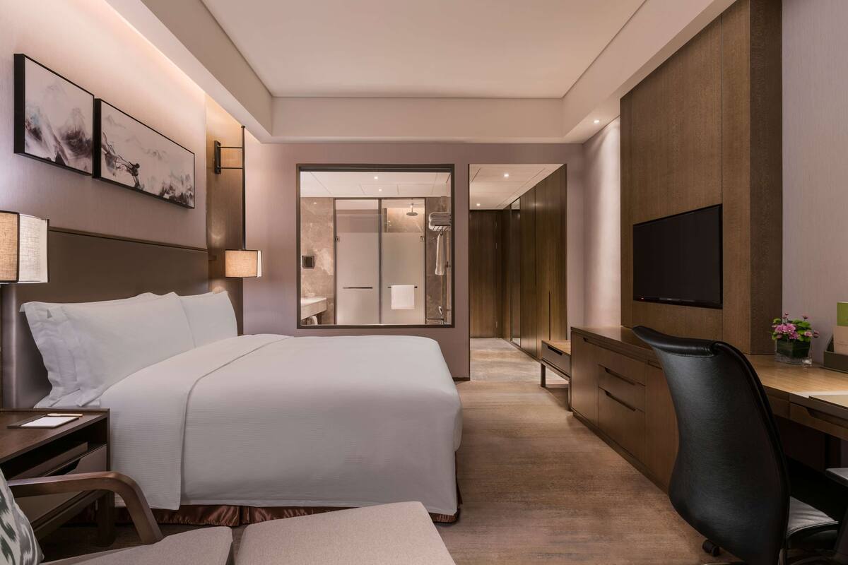 A modern hotel room with a large bed, a desk, a chair, a TV, and a bathroom visible through a glass door.