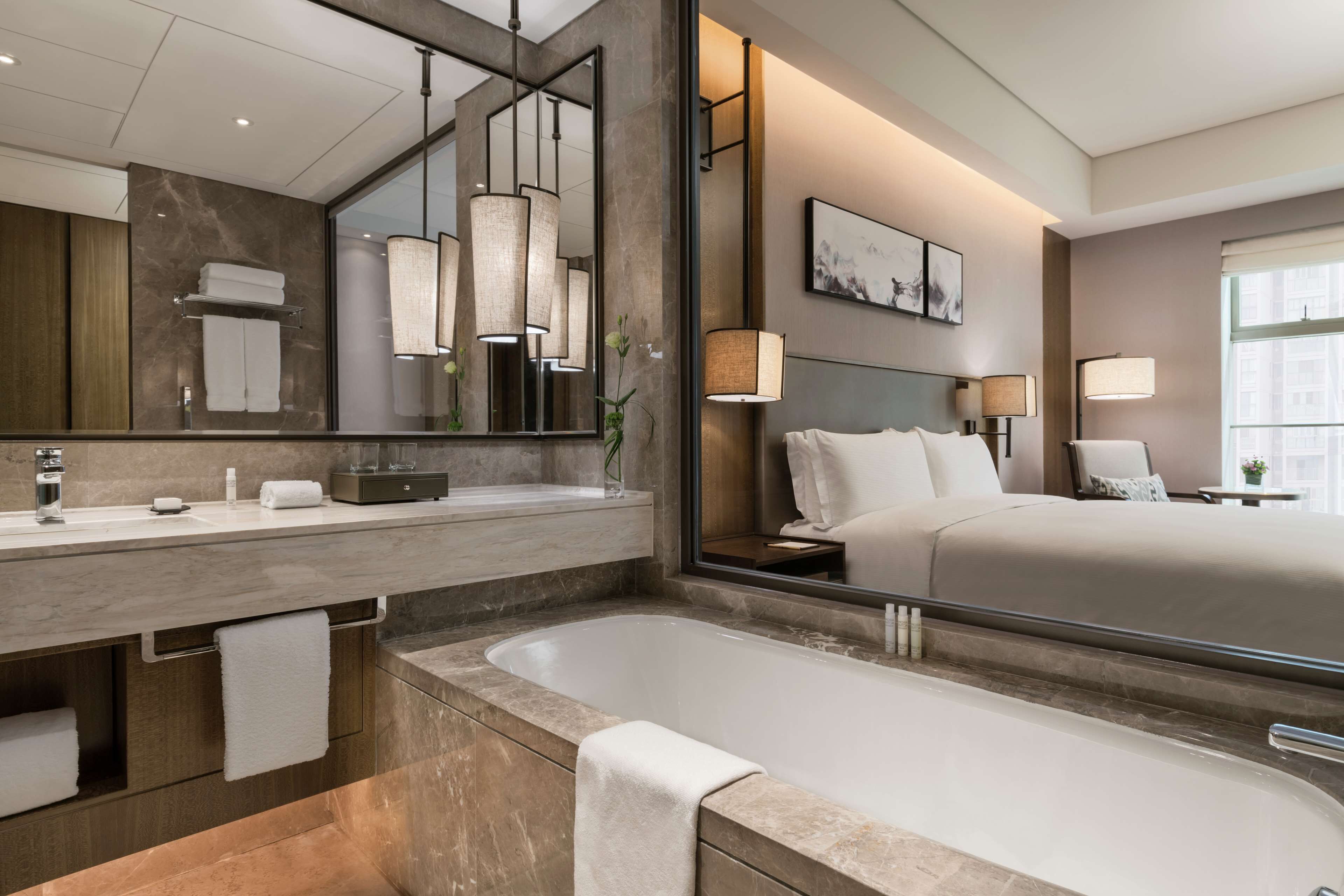 A modern hotel room with a large mirror, a bathtub, a bed with white linens, and a bedside table with a lamp.