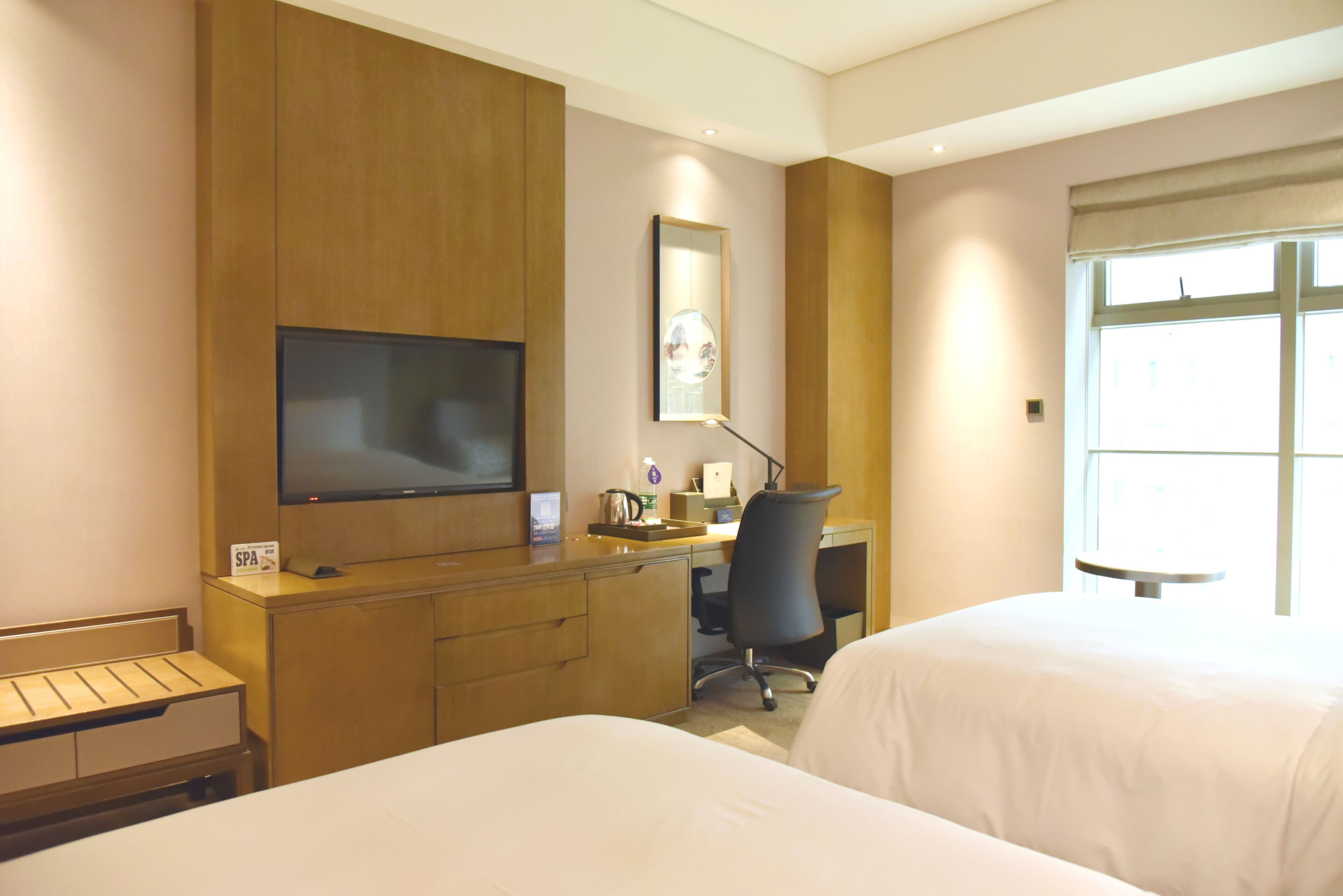 A hotel room with two beds, a desk, a TV, and a window.