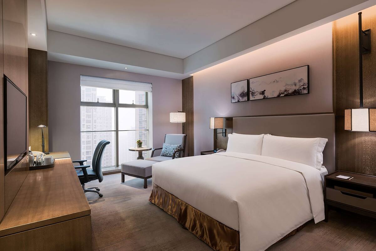 A hotel room with a large bed, a desk, a chair, a small table, and a window.