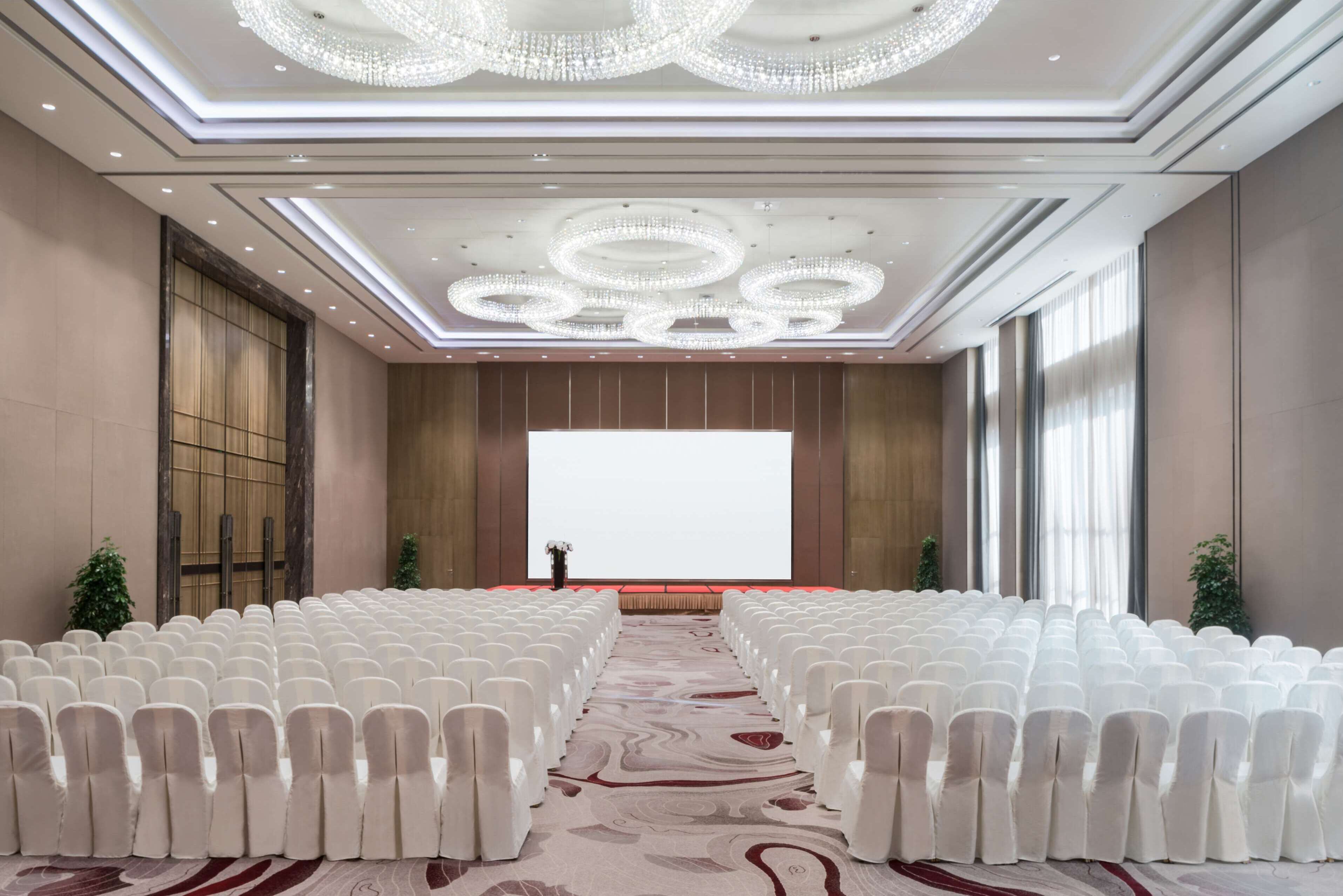 A conference room with rows of white chairs, a large screen at the front, and chandeliers on the ceiling.