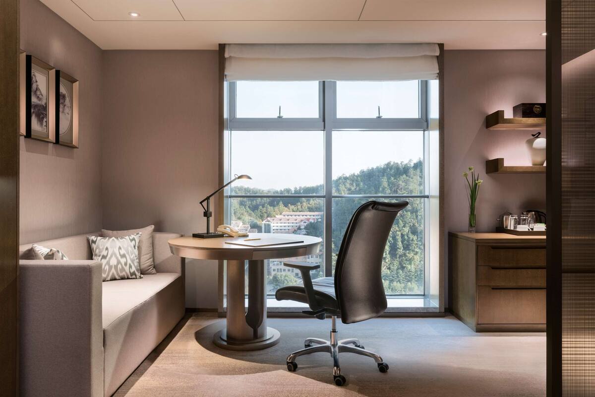 A modern office space with a round table, a black office chair, a sofa, and a large window offering a view of a forested area.