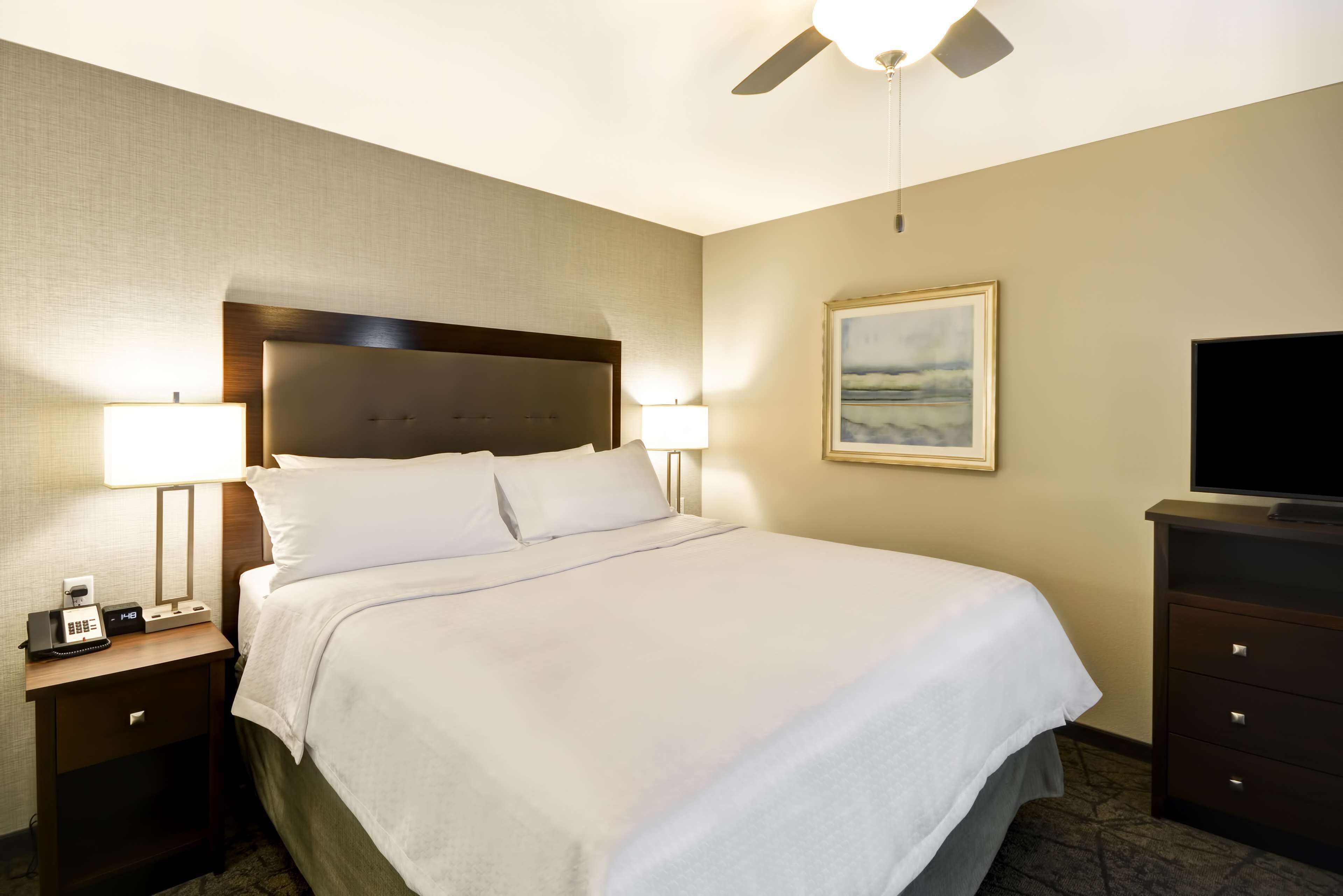 Una habitación de hotel con una cama grande, dos lámparas de noche, una mesita de noche con un teléfono, un televisor sobre un aparador y un ventilador de techo.
