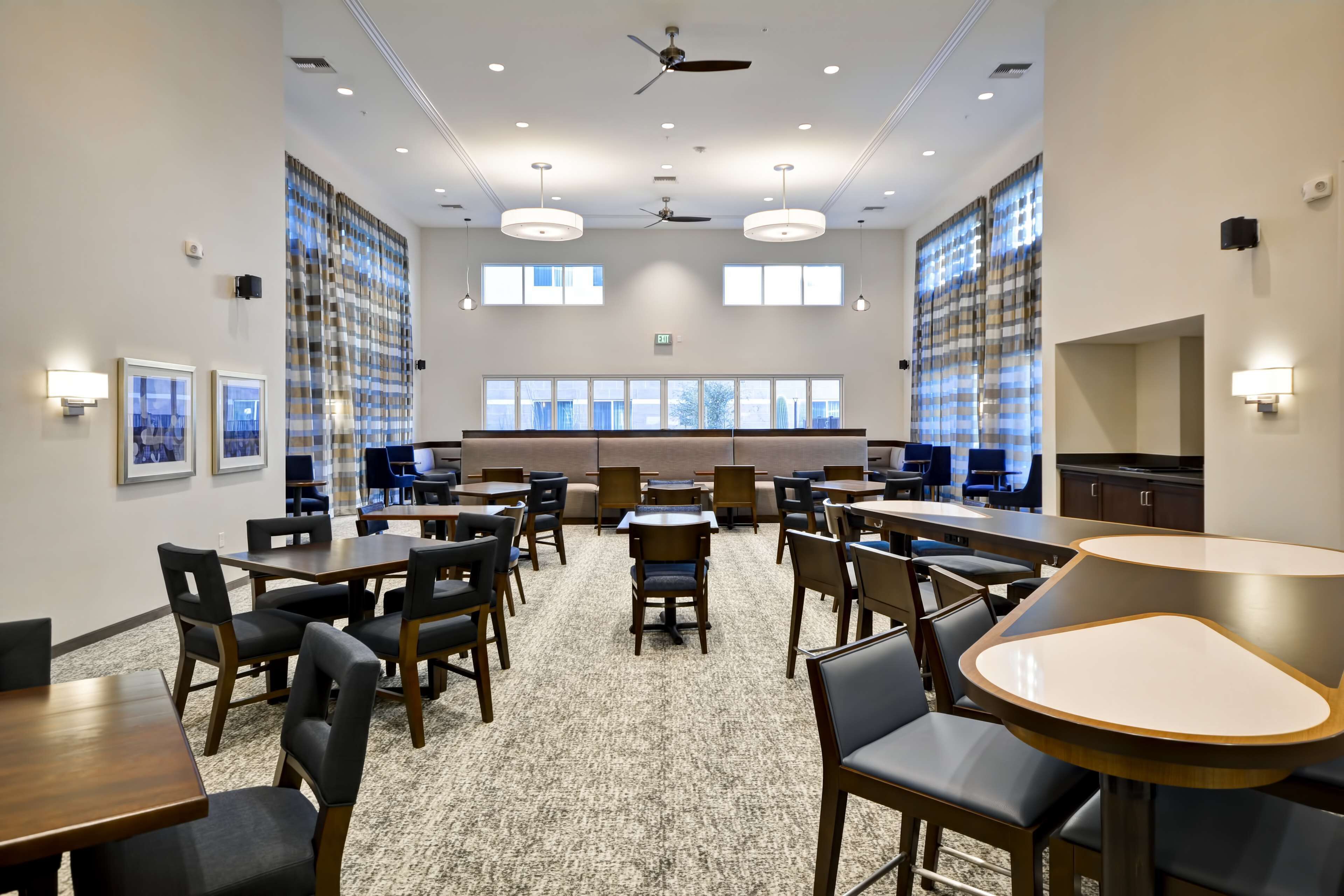 A spacious dining area with multiple tables and chairs, large windows, ceiling fans, and wall-mounted lights.