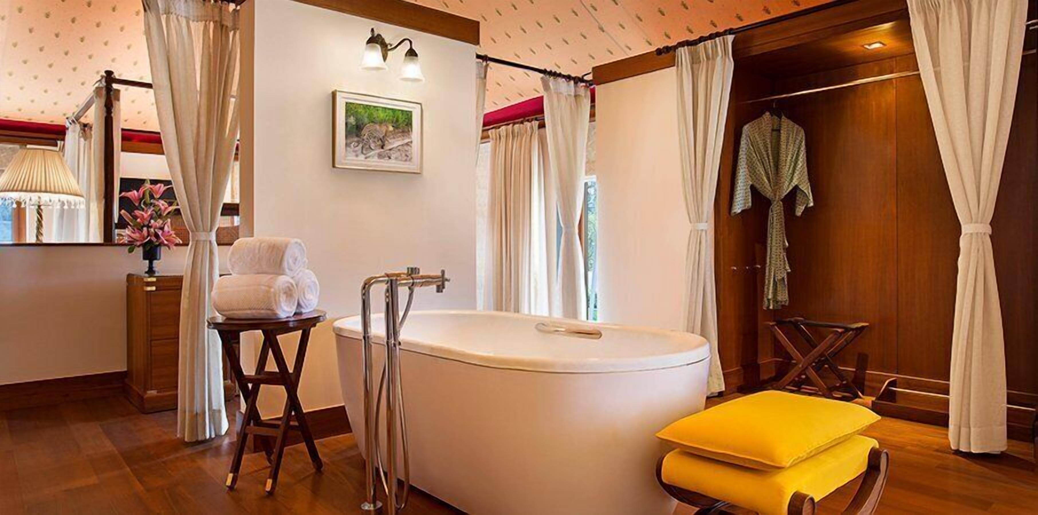 A bathroom with a bathtub, a wooden stool with towels, a wooden chair, and a yellow ottoman.