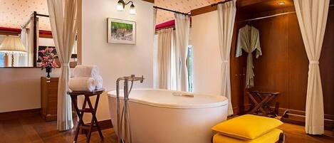A bathroom with a bathtub, a wooden stool with towels, a wooden chair, and a yellow ottoman.