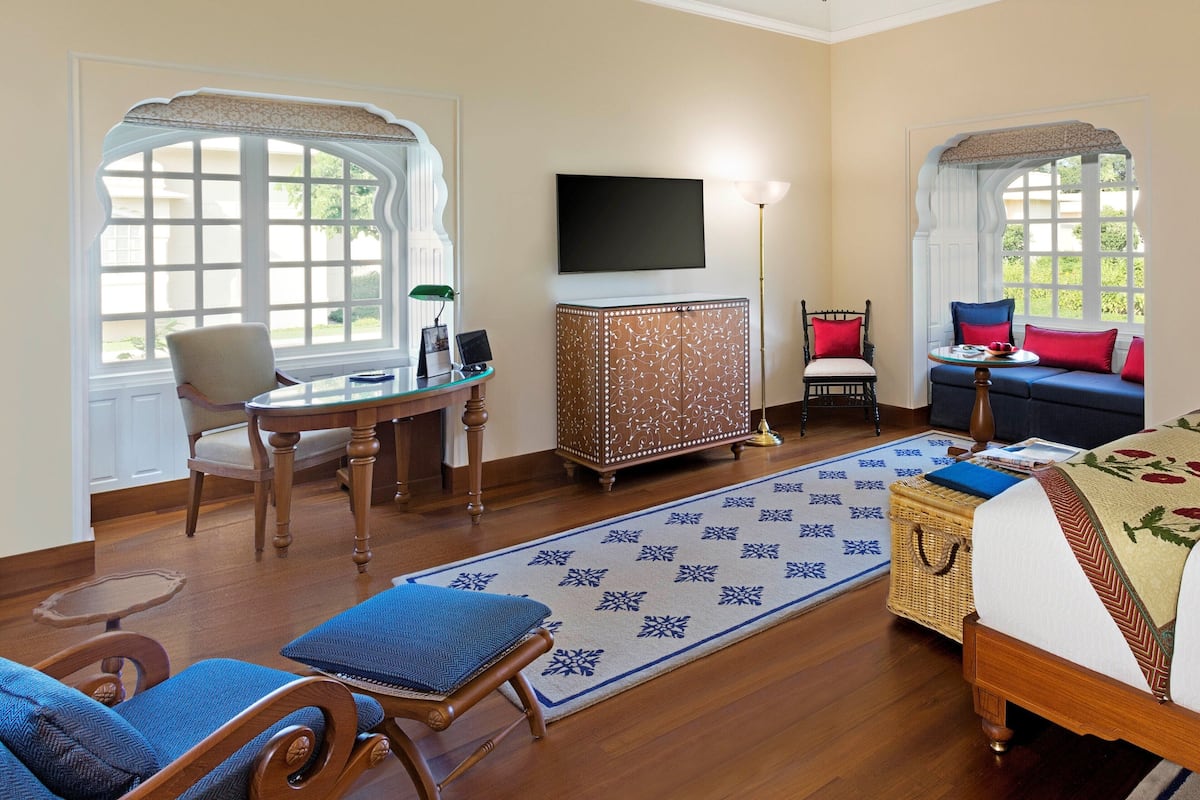 A hotel room with a wooden floor, a bed with a floral bedspread, a blue sofa, a wooden cabinet, a television mounted on the wall, and a large window with a decorative frame.