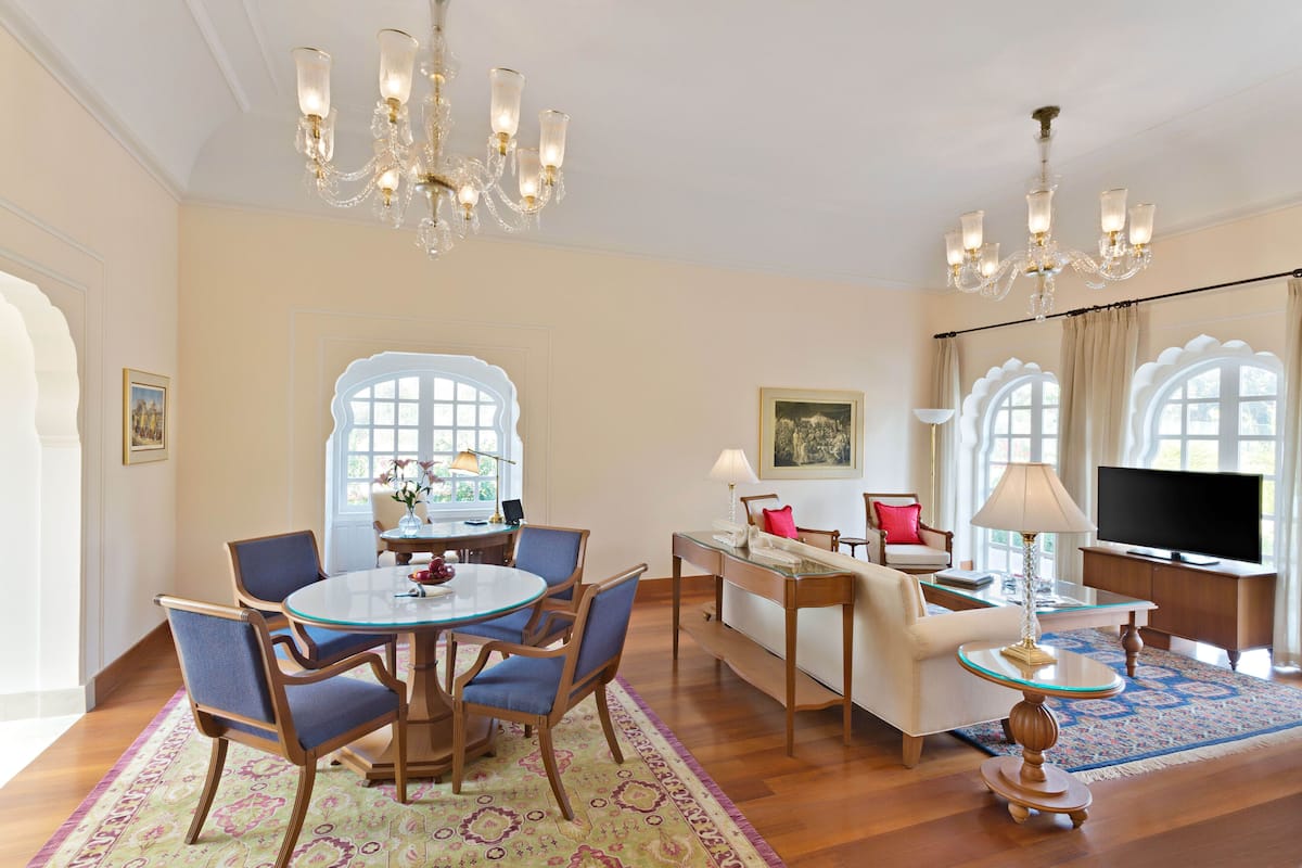A spacious living room with a round dining table, blue upholstered chairs, a sofa, a TV, and a chandelier.