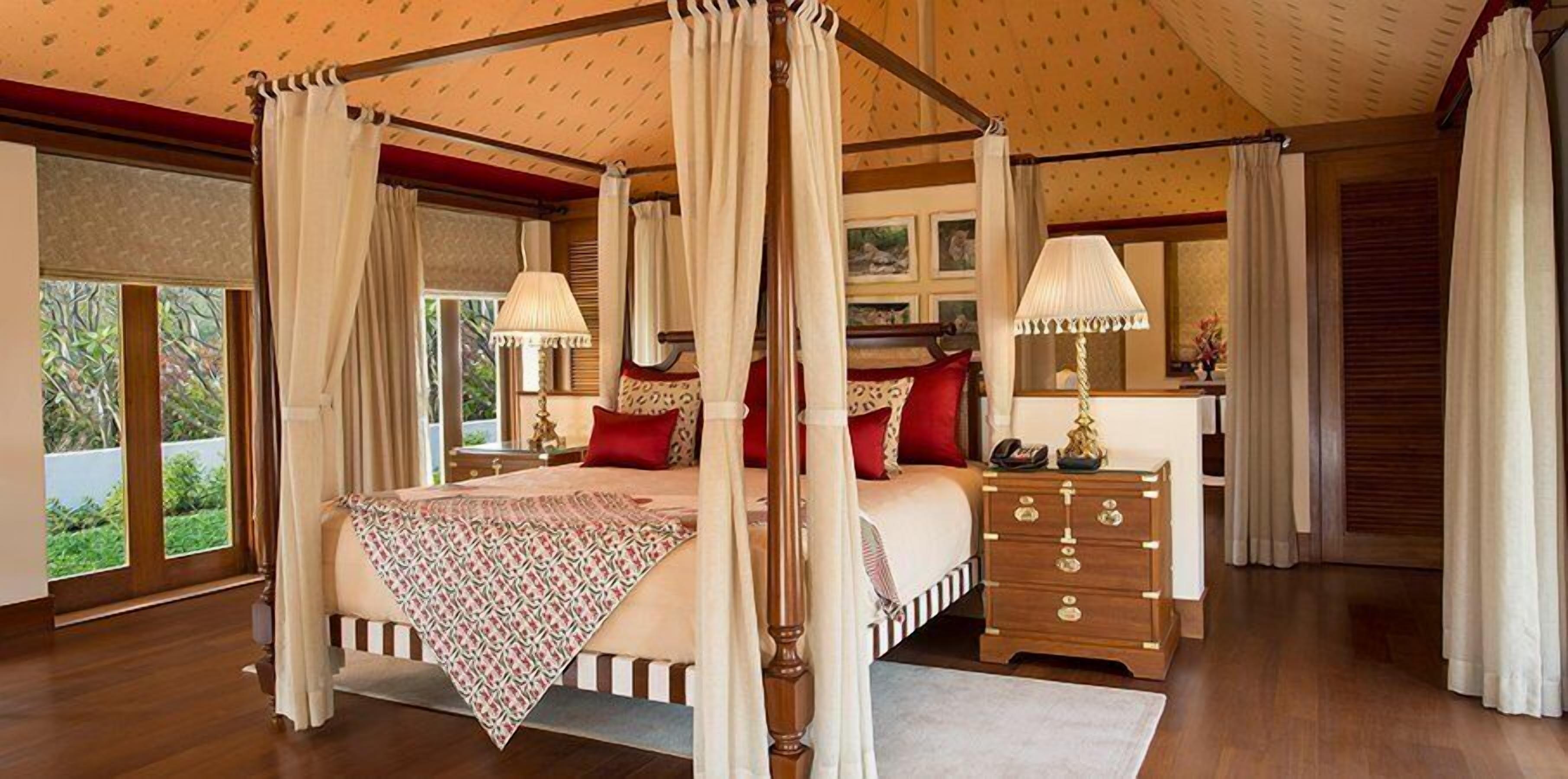 A four-poster canopy bed with a patterned throw, wooden nightstands, and a view of greenery outside.