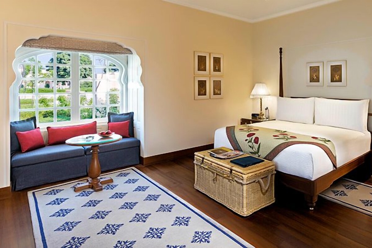 A bedroom with a bed, a sofa, a nightstand, a lamp, a dresser, and a window with a view.