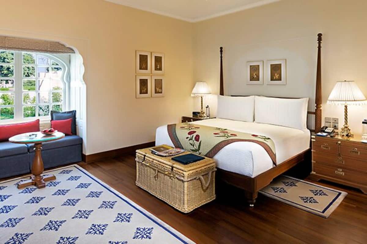 A bedroom with a bed, a sofa, a nightstand, a lamp, a dresser, and a window with a view.