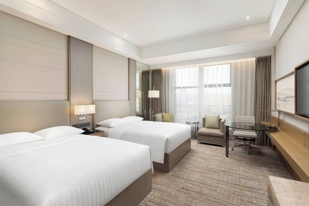 A hotel room with two beds, a desk, a chair, a TV, and a large window with curtains.