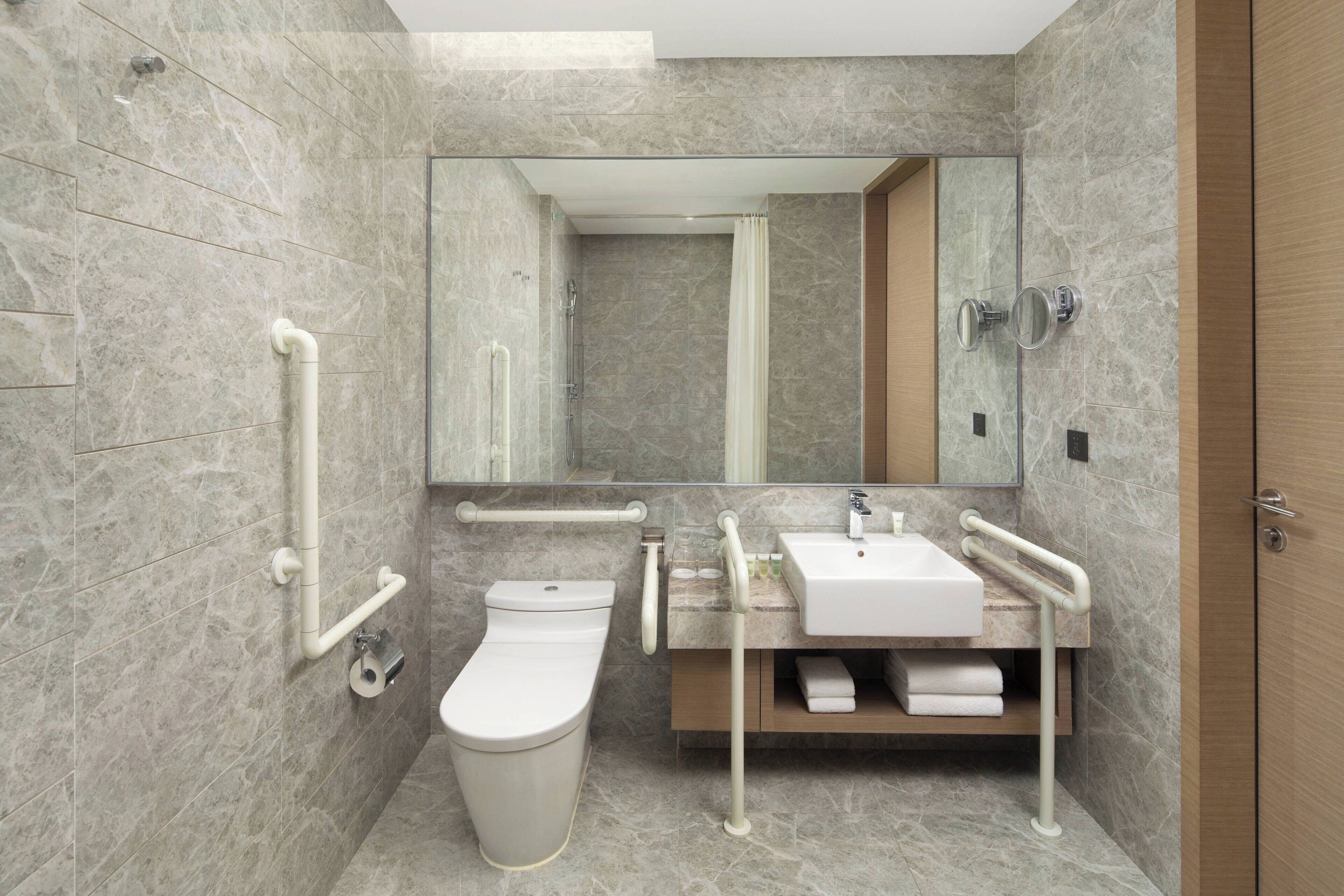 Un baño moderno con inodoro, lavamanos y espejo. Cuenta con barras de apoyo, un perchero y una zona de ducha.