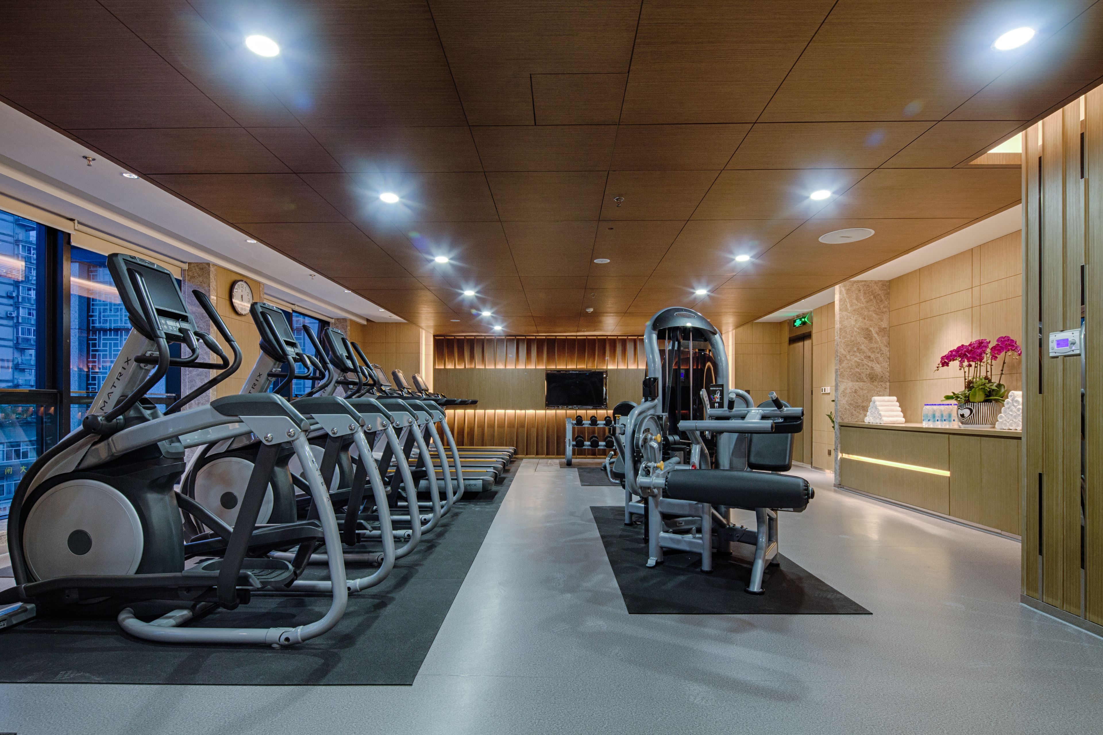 Un gimnasio con cintas de correr, máquinas de pesas y una recepción con un reloj y una planta.