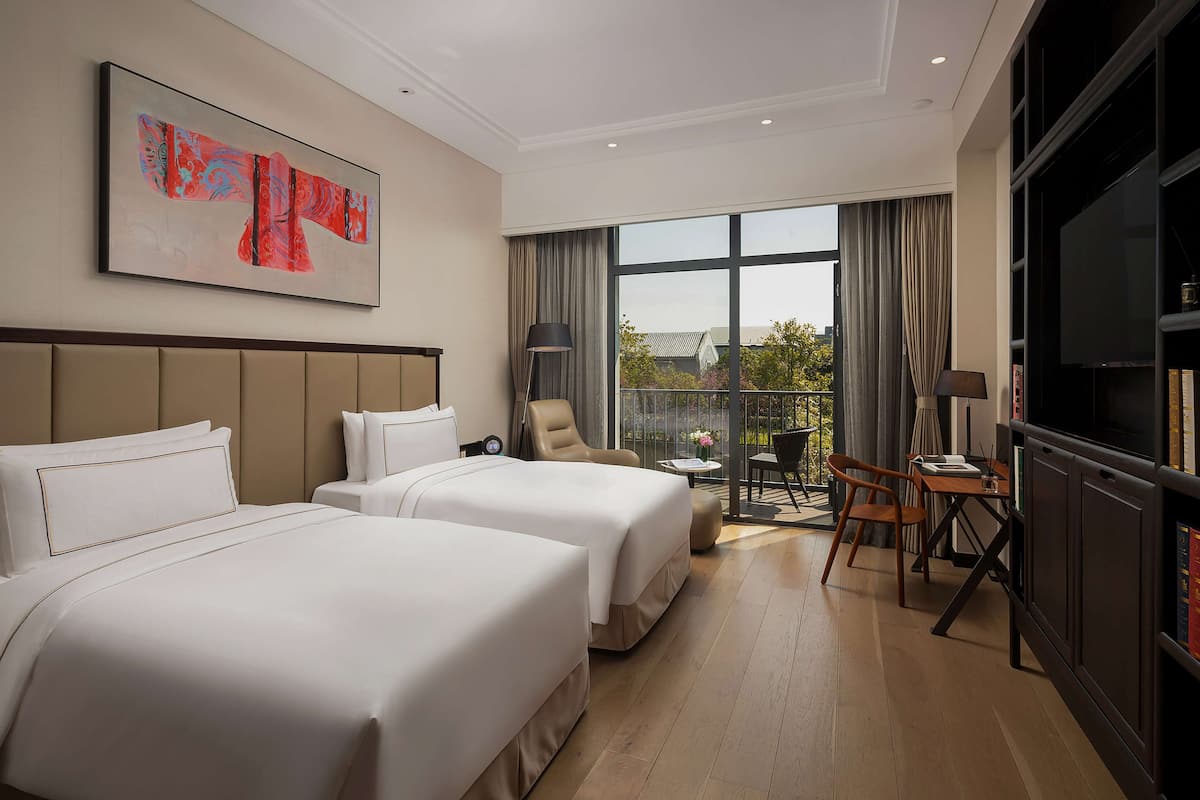 A hotel room with two beds, a TV, a desk, and a balcony with a view.