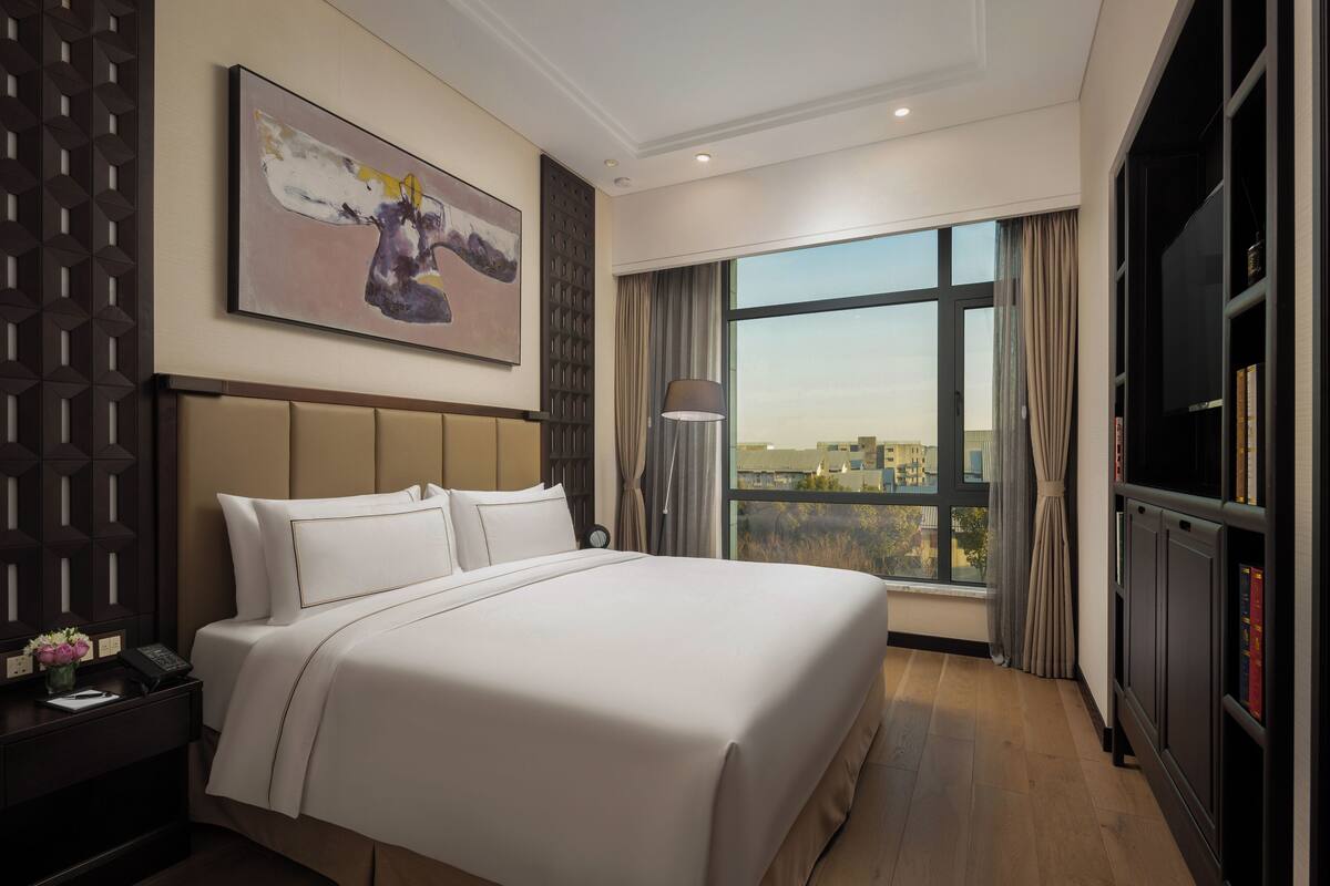 A hotel room with a large bed, a painting on the wall, a nightstand with a lamp, and a view of the city through large windows.
