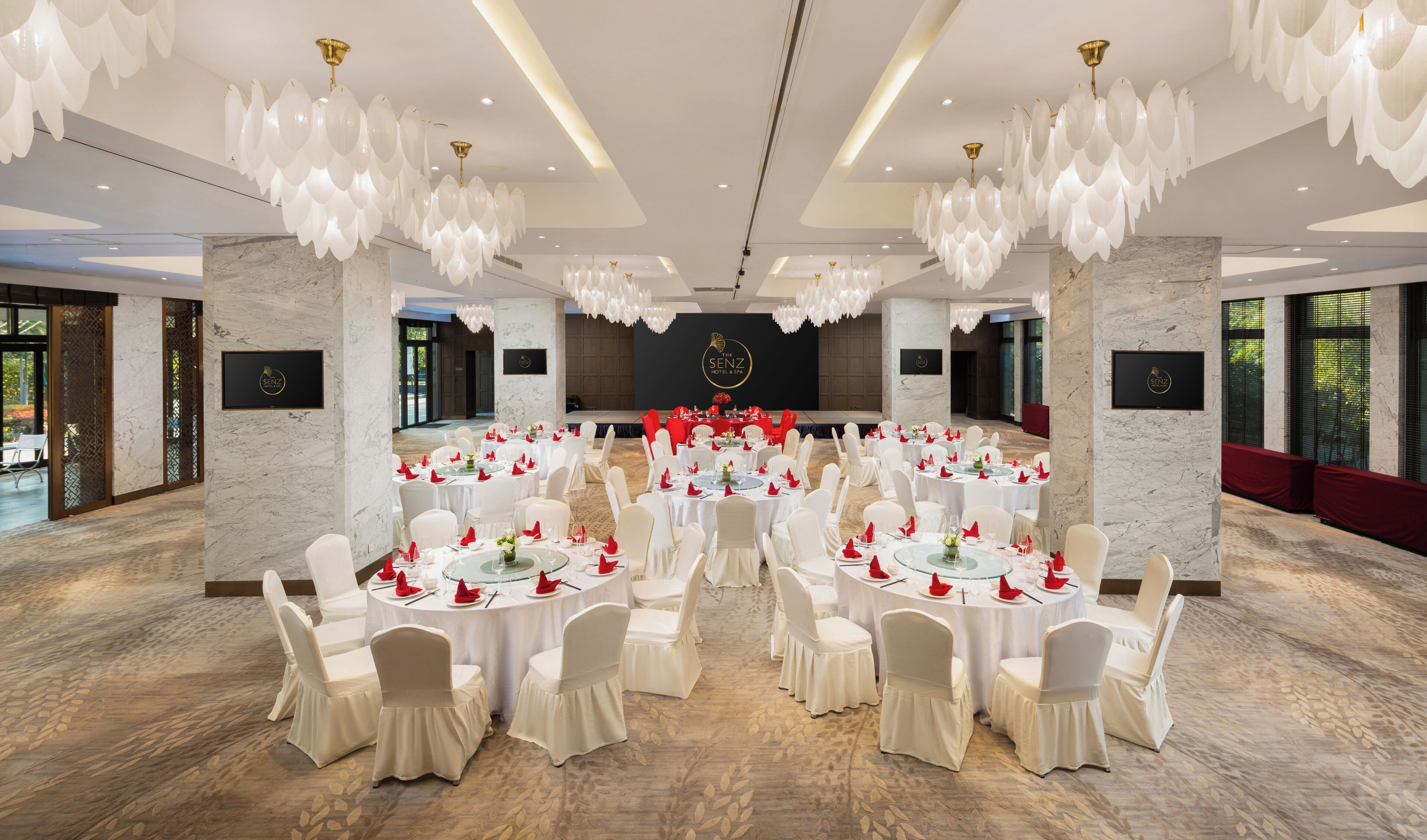 A banquet hall with round tables, white chairs, chandeliers, and a stage with a logo.