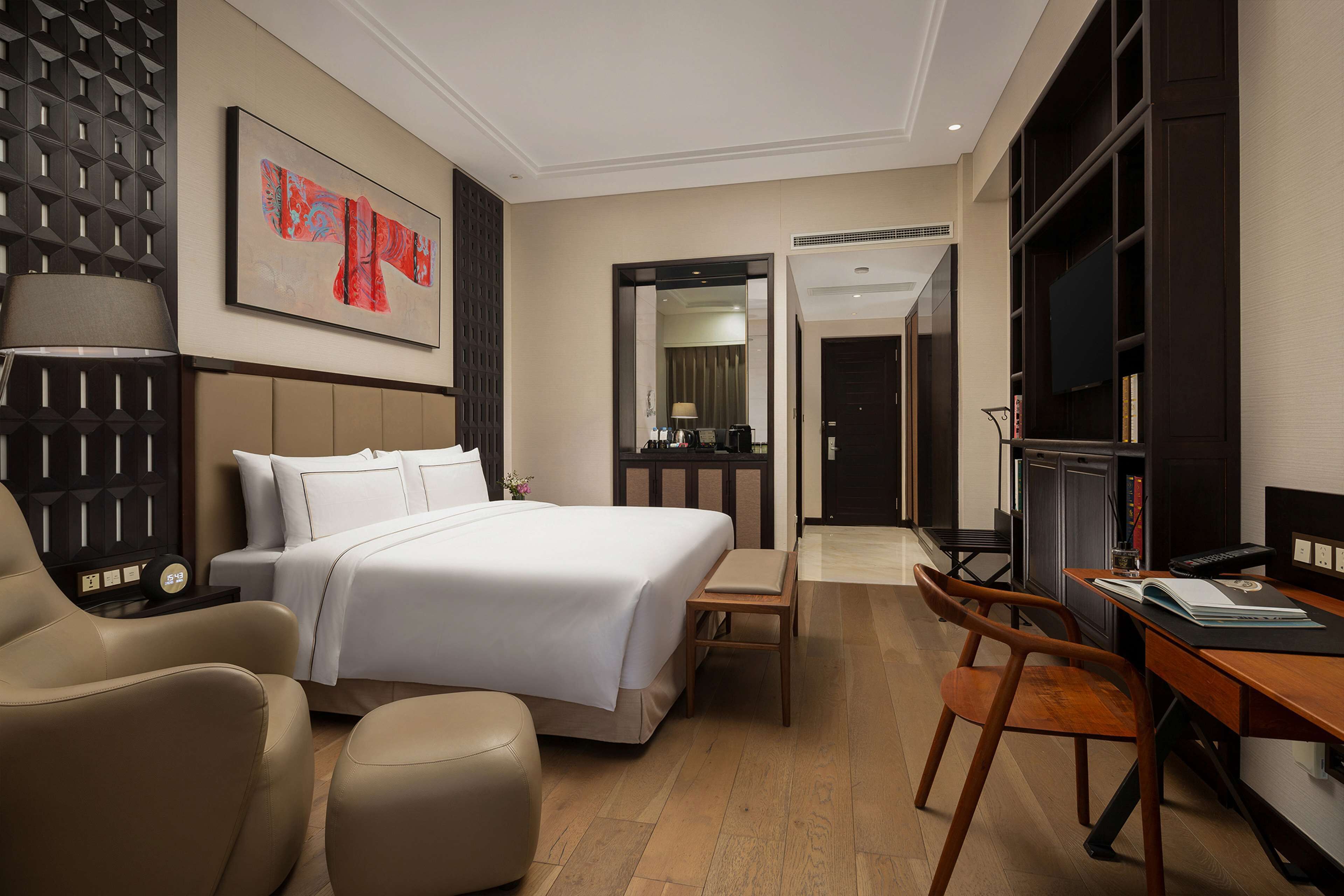 A hotel room with a bed, a desk, a chair, a TV, and a bookshelf.