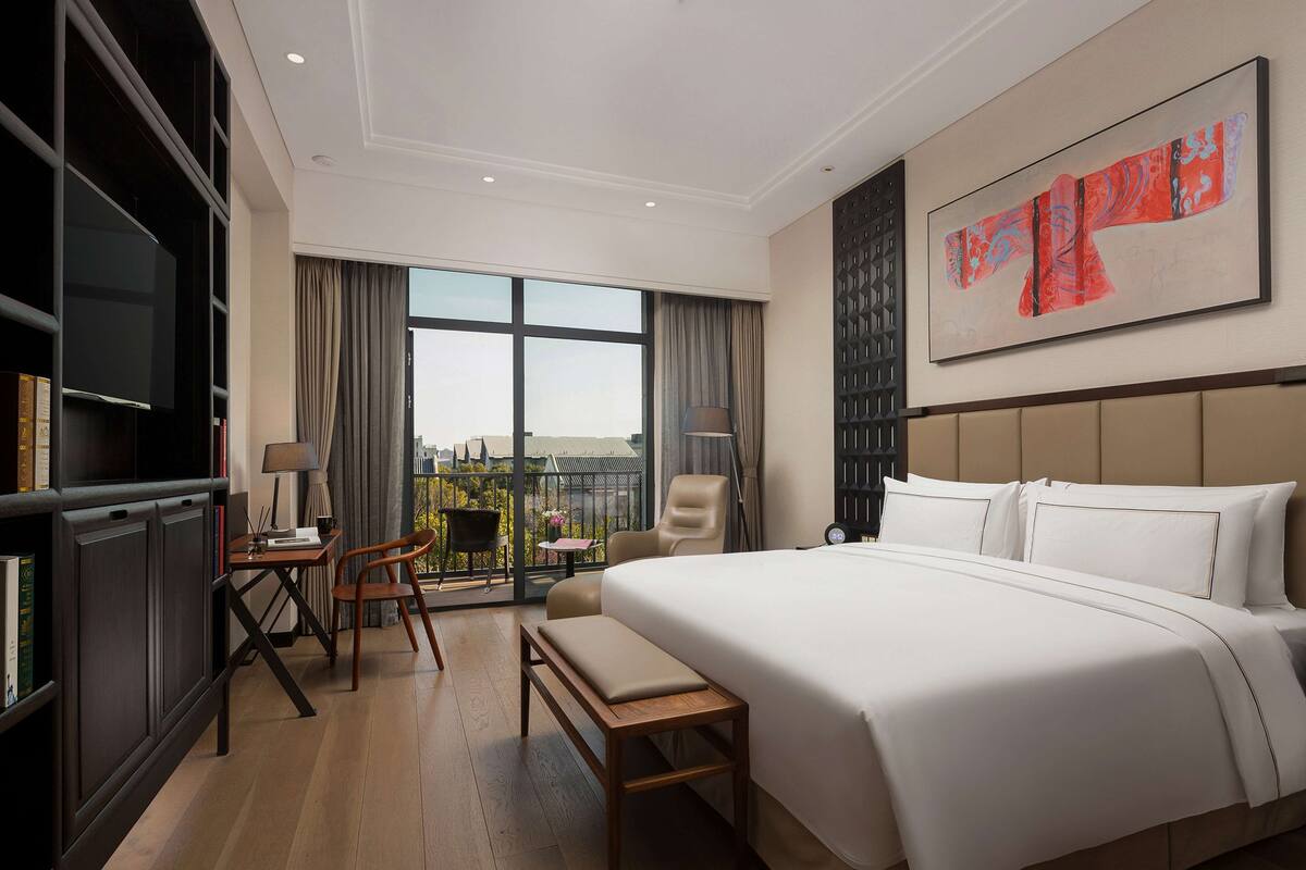 A hotel room with a large bed, a desk with a chair, a television, and a balcony with a view.
