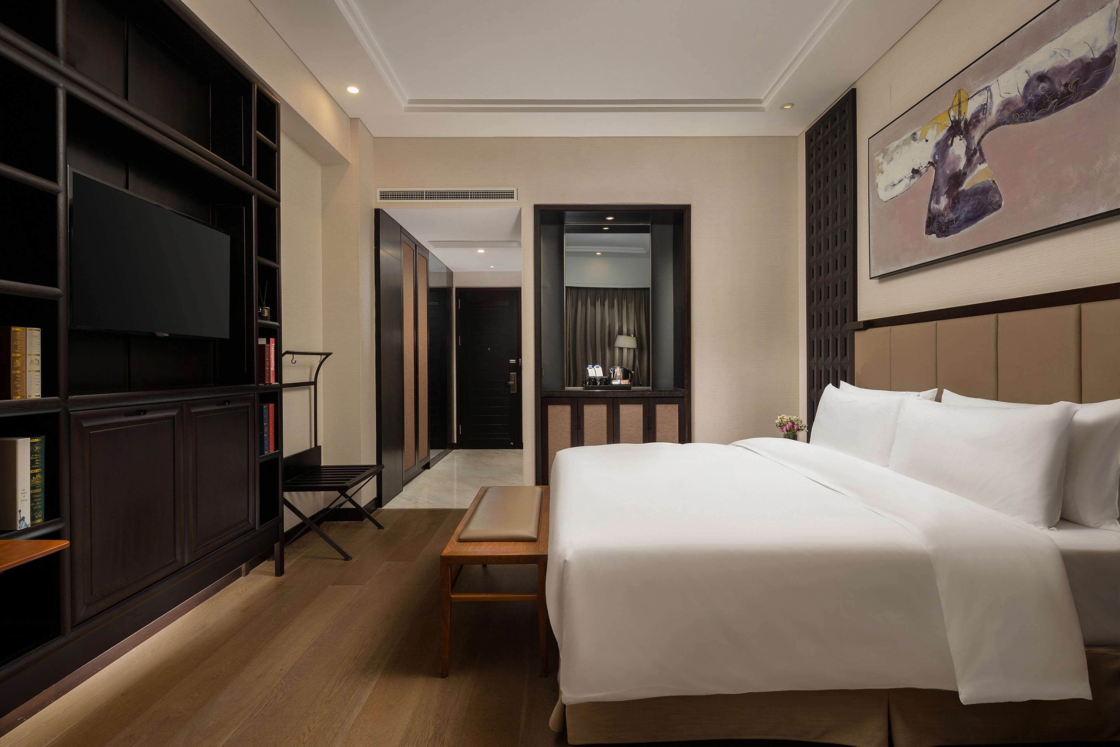 A hotel room with a large bed, a TV mounted on the wall, a bookshelf, and a painting.