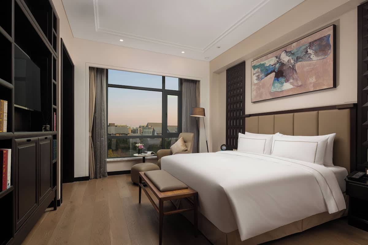 A hotel room with a large bed, a desk, a chair, a bookshelf, and a large window with a city view.