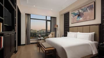 A hotel room with a large bed, a desk, a chair, a bookshelf, and a large window with a city view.