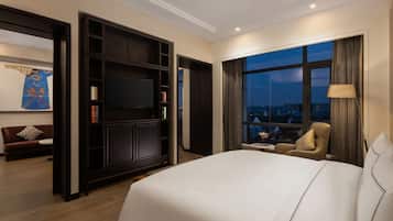 A hotel room with a large bed, a TV, a bookshelf, a sofa, and a view of the city at night.