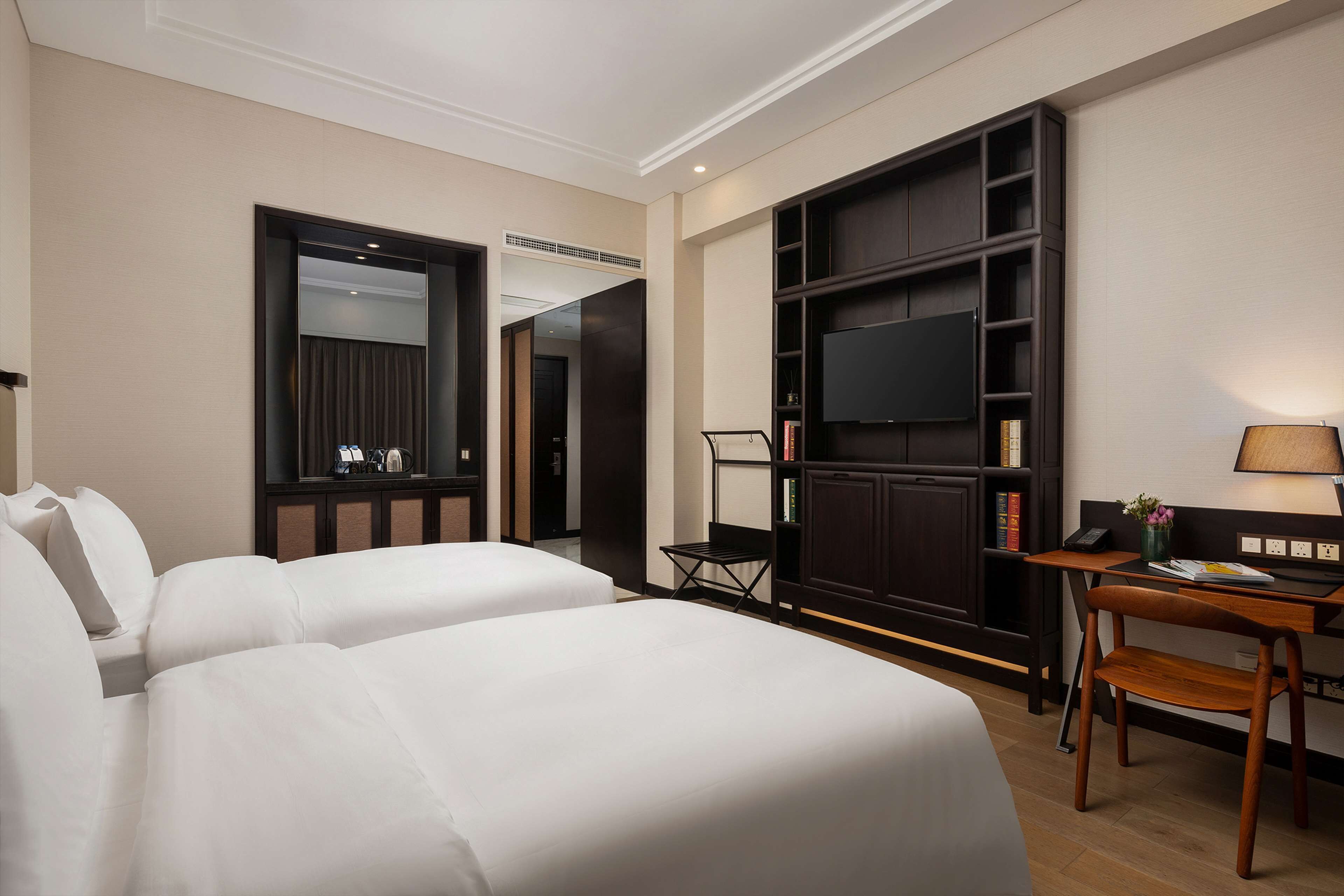 A hotel room with two beds, a desk, a chair, a TV, and a bookshelf.
