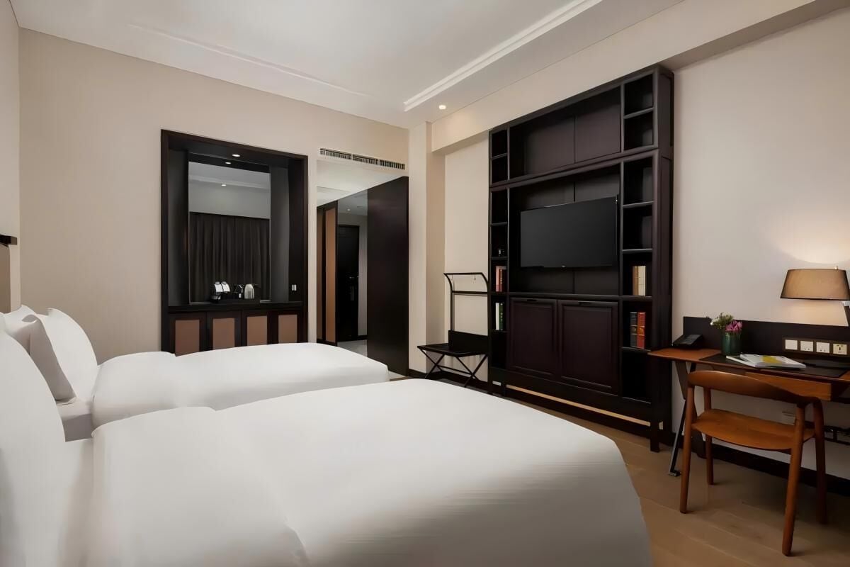 Een hotelkamer met twee bedden, een bureau met stoel, een televisie en een boekenplank.