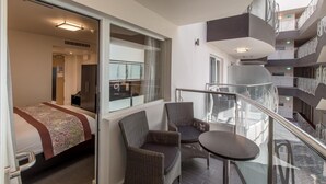 Quarto de hotel moderno com varanda, uma cama com colcha florida, cadeiras de vime e uma pequena mesa.