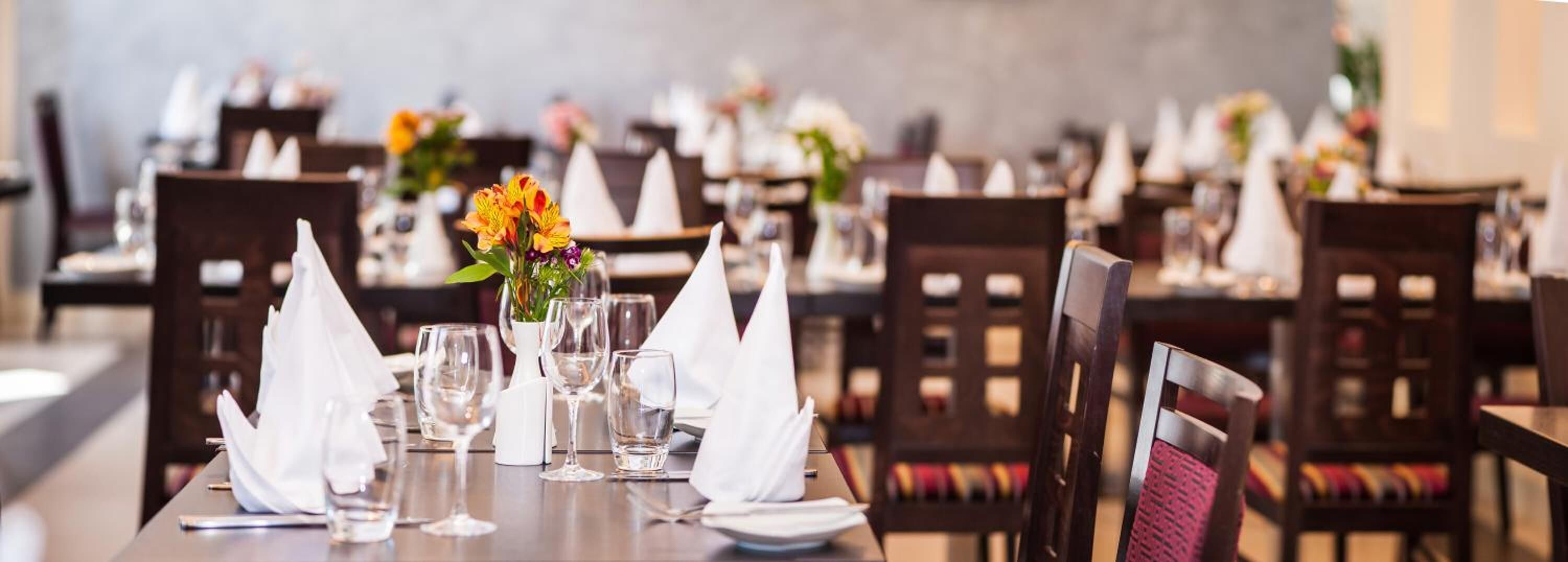 Un restaurante con mesas ordenadas, cada una con servilletas blancas, vasos transparentes y centros de mesa florales.