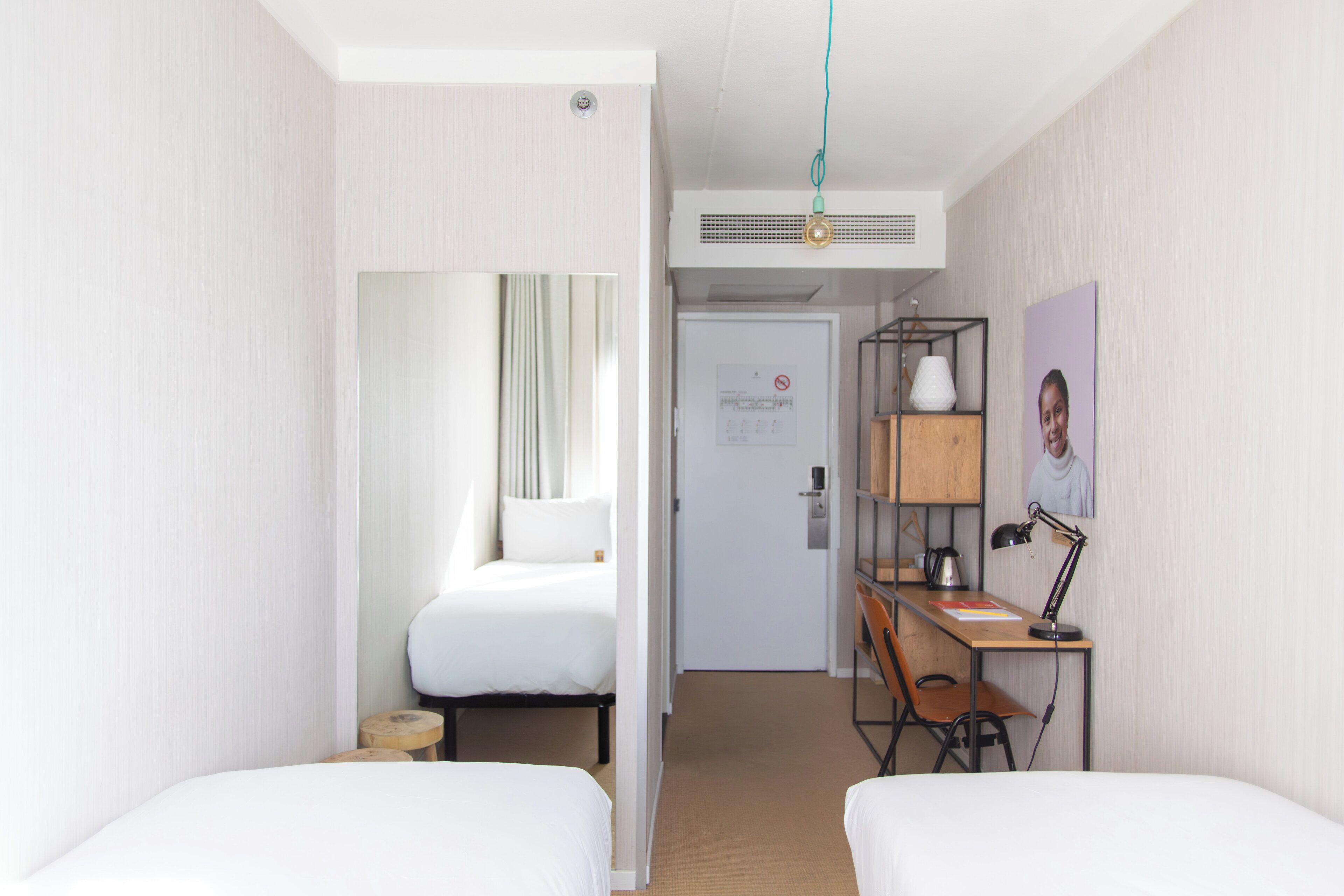 Une chambre d’hôtel avec deux lits, un bureau avec une chaise, une lampe, une étagère avec un tableau, et une porte donnant sur une autre pièce.