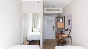 Une chambre d’hôtel avec deux lits, un bureau avec une chaise, une lampe, une étagère avec un tableau, et une porte donnant sur une autre pièce.