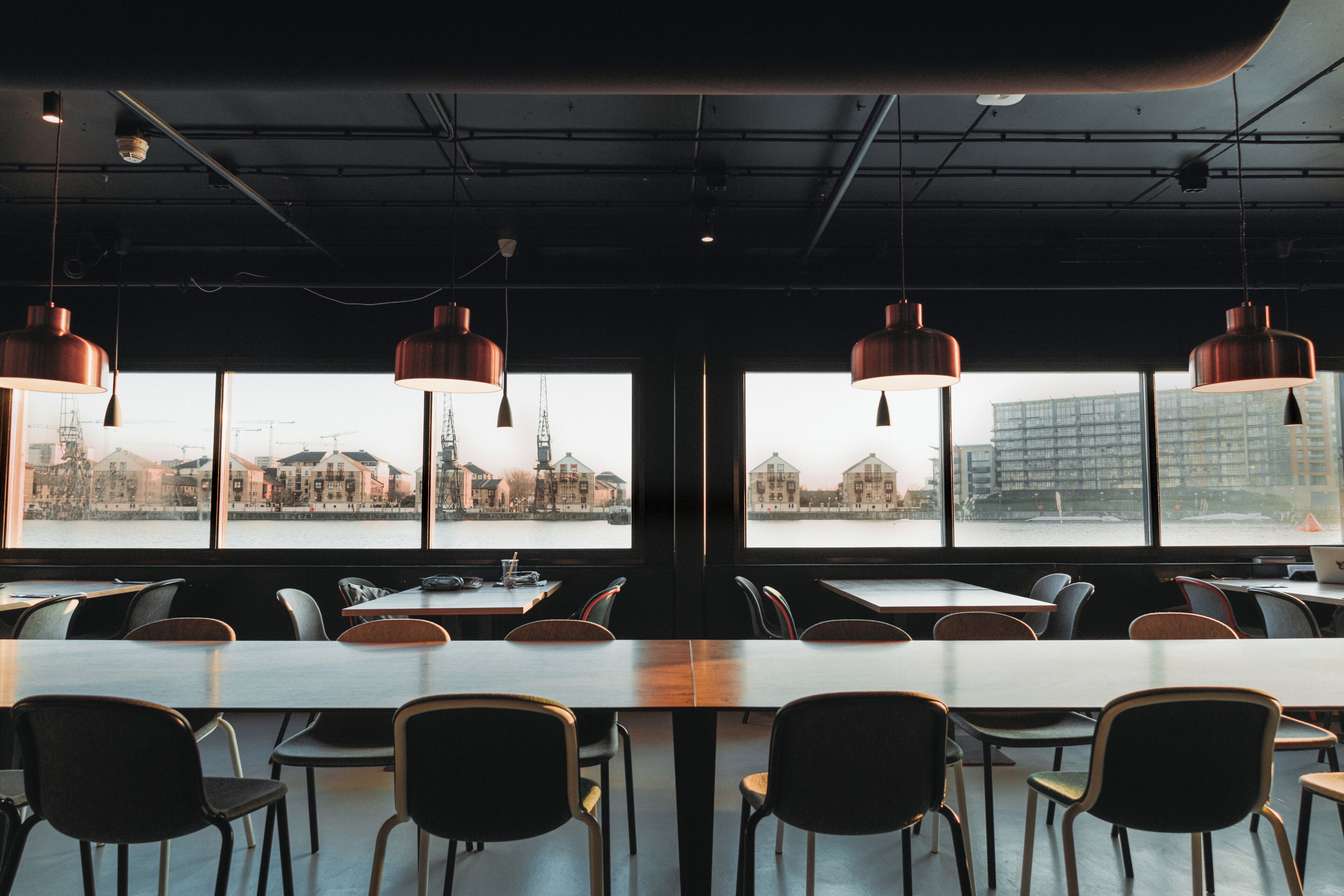A modern dining area with long tables and chairs, large windows offering views of a cityscape and buildings.