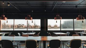 A modern dining area with long tables and chairs, large windows offering views of a cityscape and buildings.