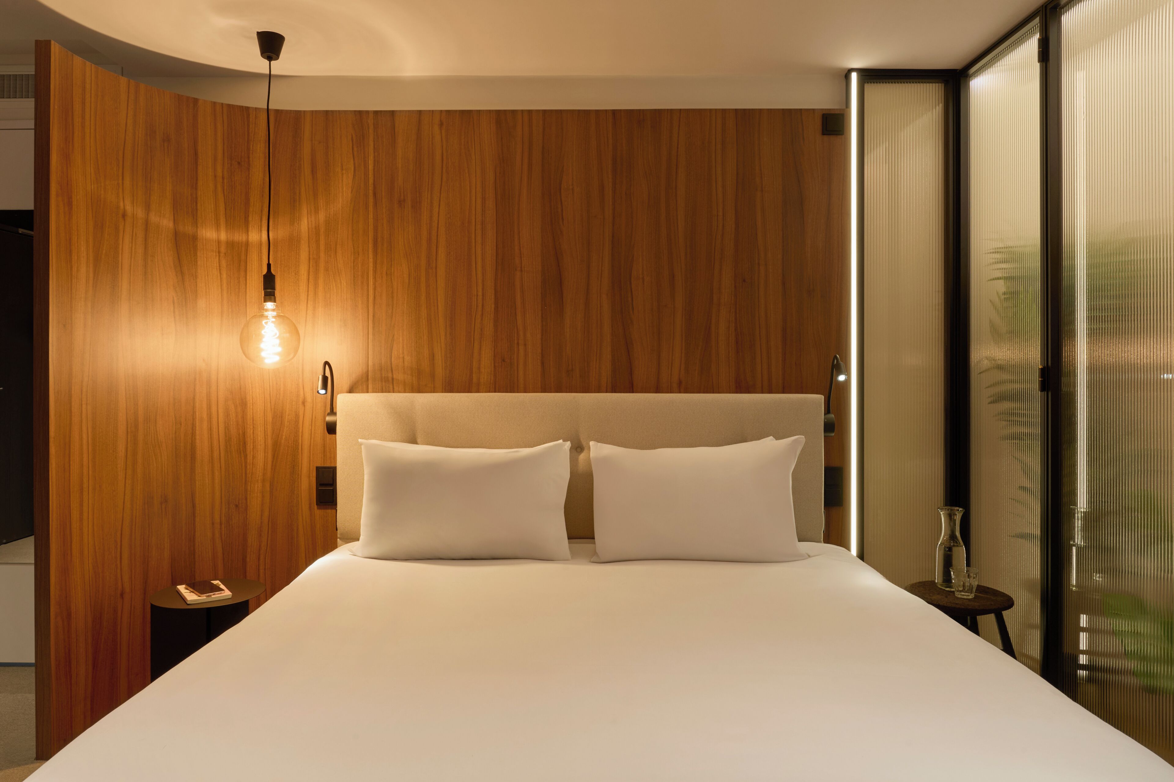 A modern hotel room with a wooden headboard, a single bed with white linens, a bedside table with a lamp, and a view through a glass door.