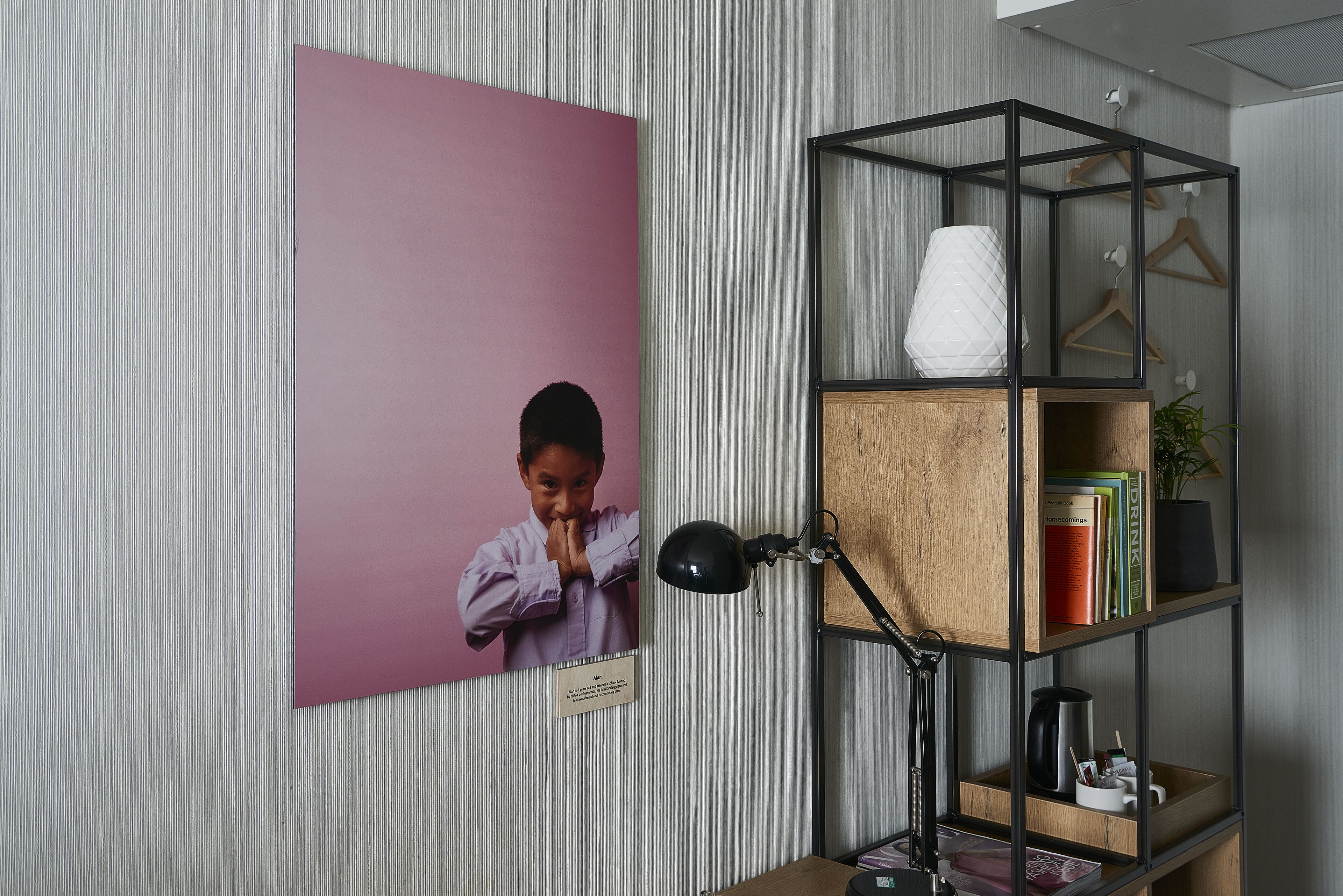 Een kamer met een lijst eraan de muur, een bureau met een lamp, en een houten boekenkast met boeken en een vaas.