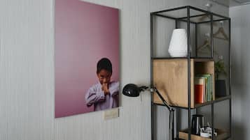 Een kamer met een lijst eraan de muur, een bureau met een lamp, en een houten boekenkast met boeken en een vaas.
