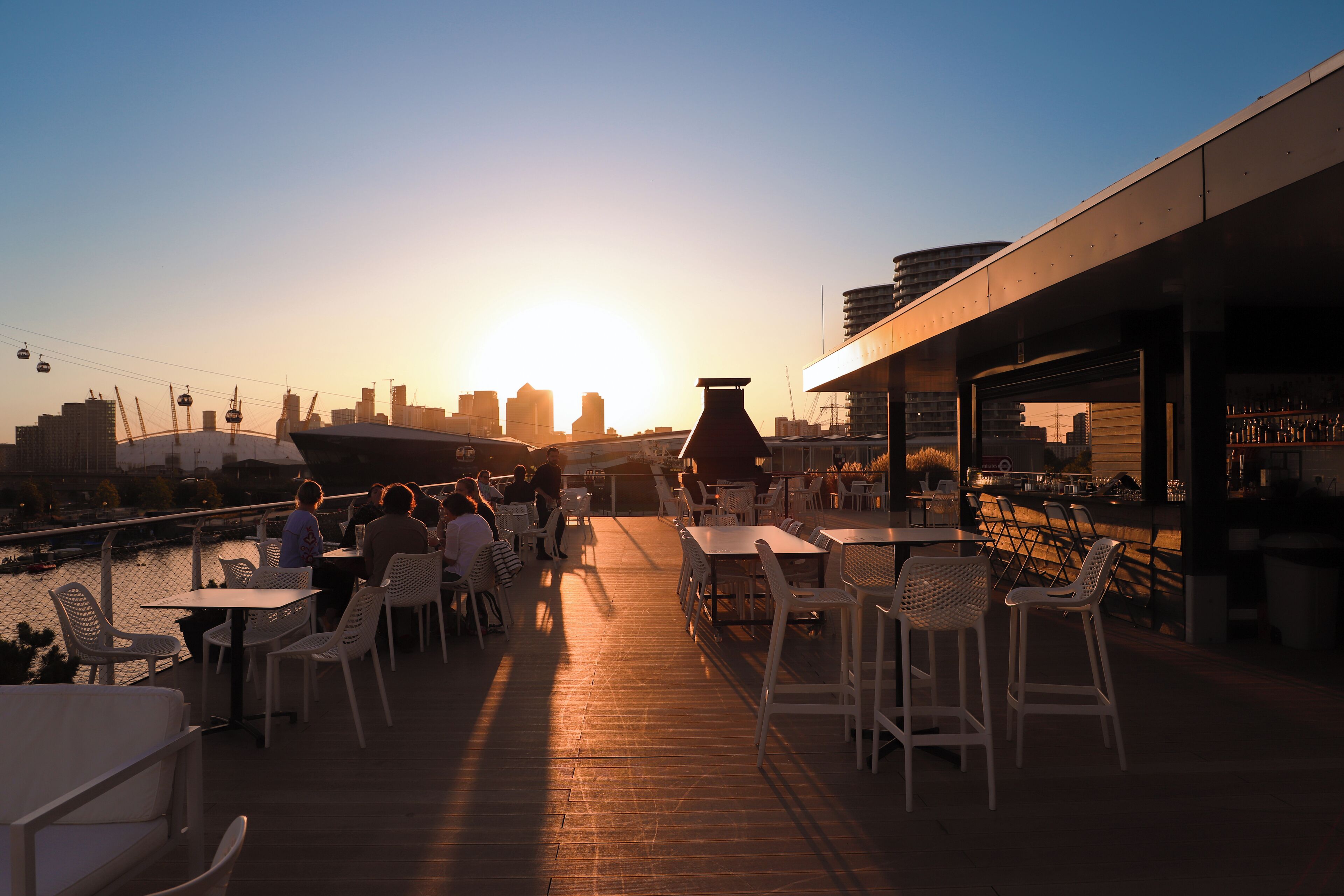 A rooftop bar with tables and chairs, overlooking a city skyline at sunset.