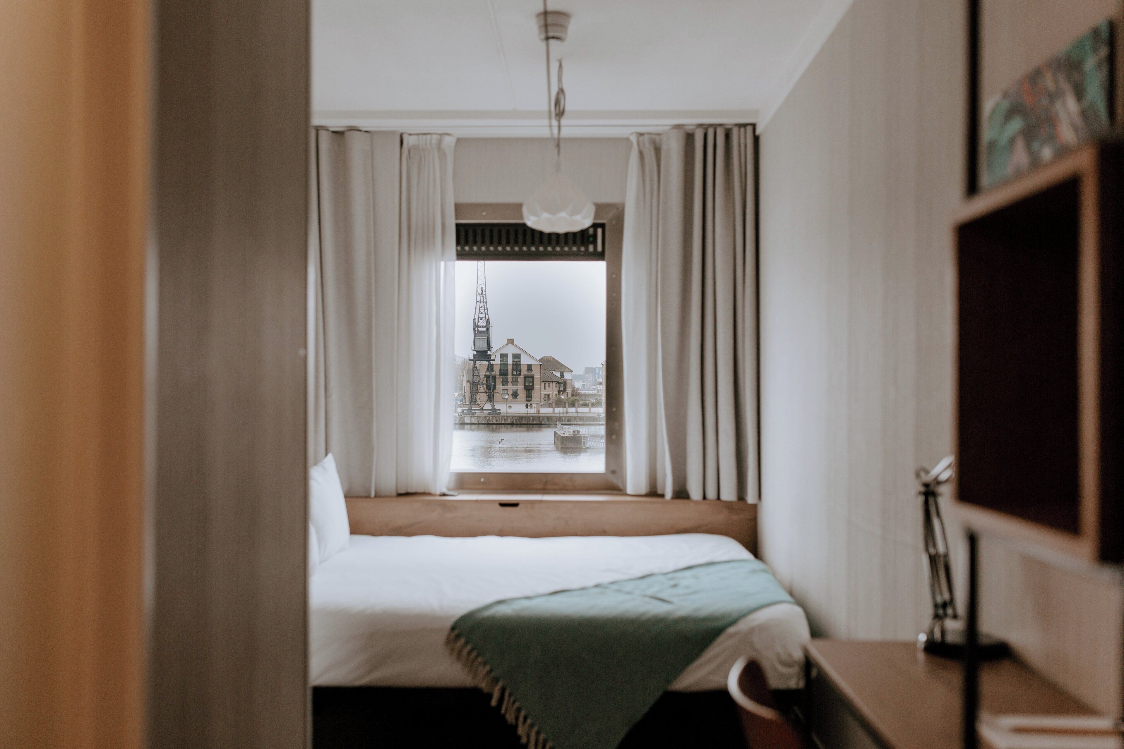 Une chambre d’hôtel avec un lit, une fenêtre avec des rideaux, une vue sur un immeuble et une étagère avec une lampe.