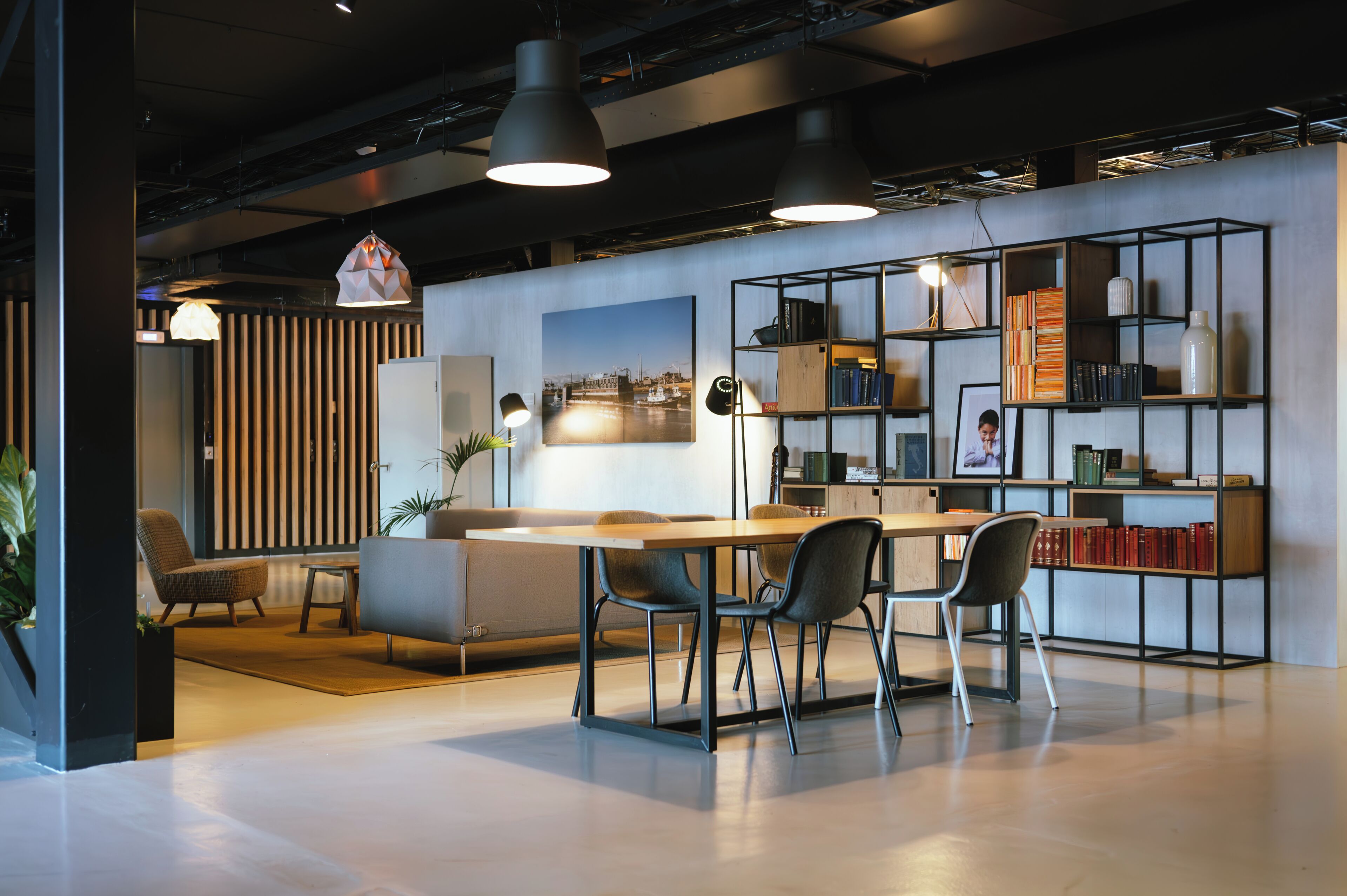 A modern office space with a wooden desk, chairs, a bookshelf, and a large window with blinds.