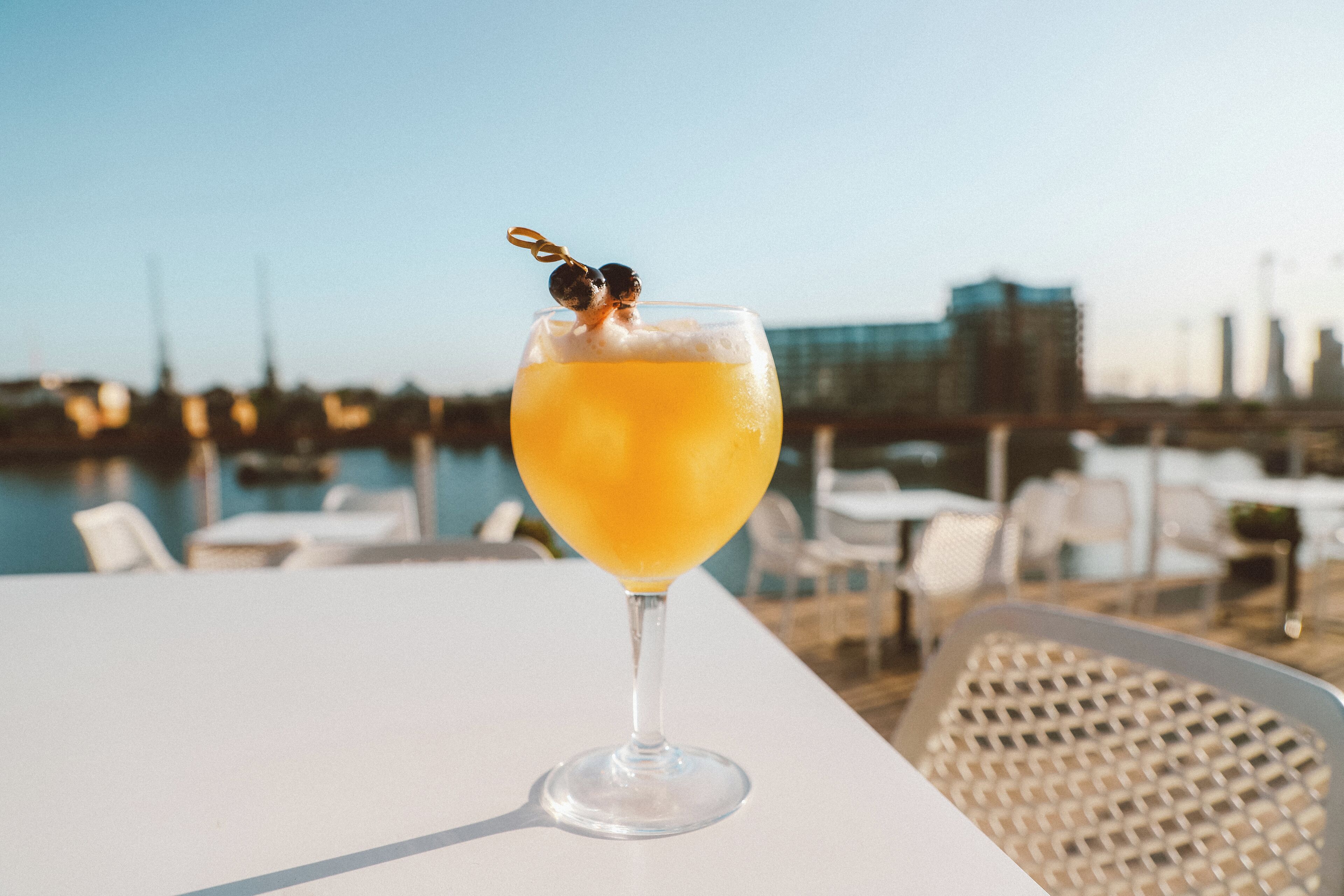 Ein Cocktailglas mit einer Garnitur auf einem Tisch am Ufer, im Hintergrund Stühle und Tische.