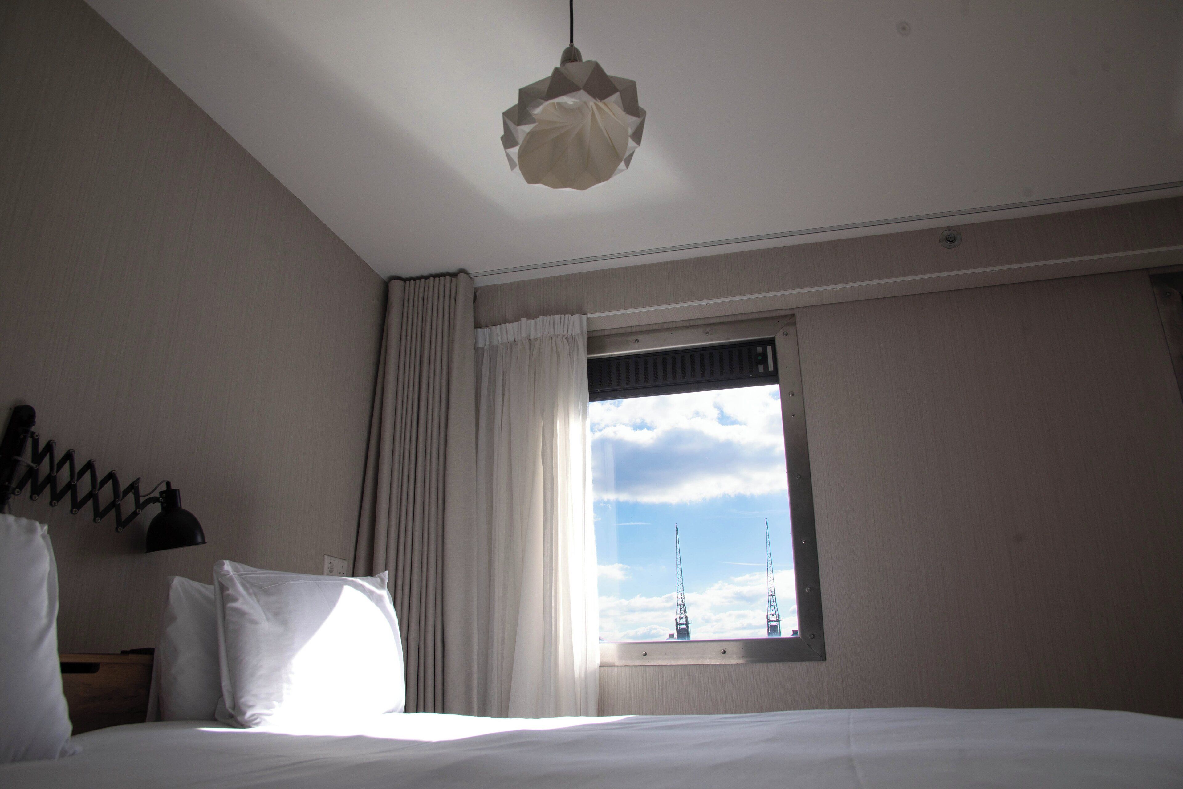 A hotel room with a bed, a window with curtains, a hanging lamp, and a view of the sky.