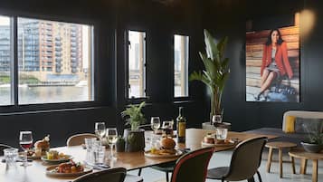 Un área de comedor con vista a un edificio al otro lado del agua, una mesa dispuesta con comida y bebidas, y una planta grande en maceta.
