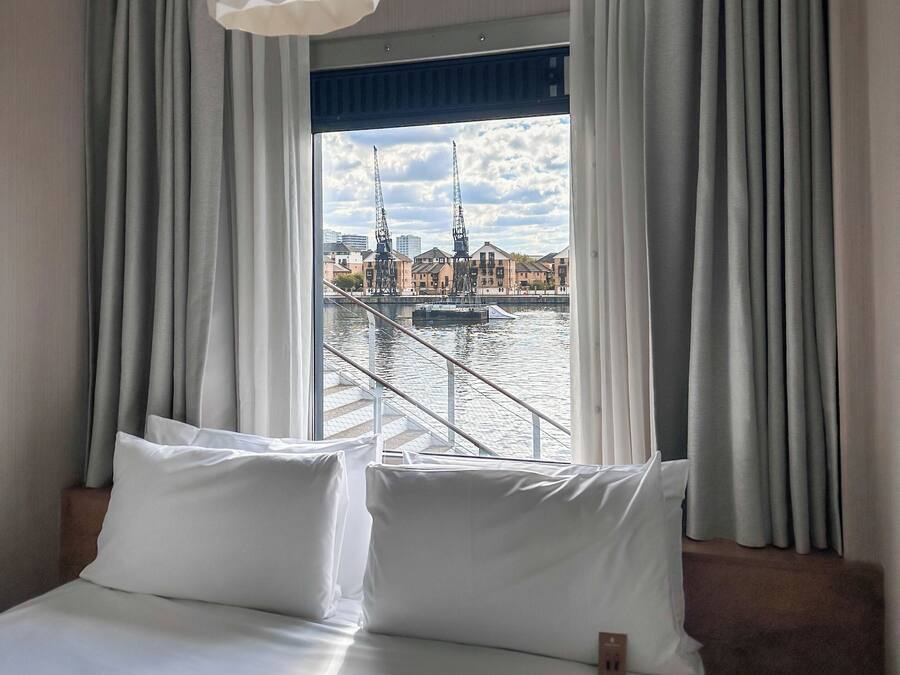 Quarto de hotel com uma cama grande, roupa de cama branca e vista para um rio com edifícios e guindastes.