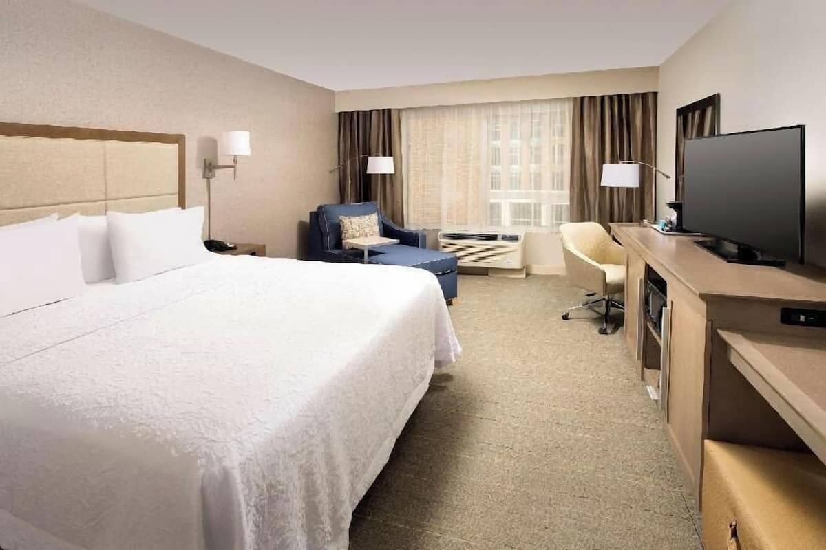 A hotel room with a large bed, a desk with a flat-screen TV, a chair, and a desk lamp.
