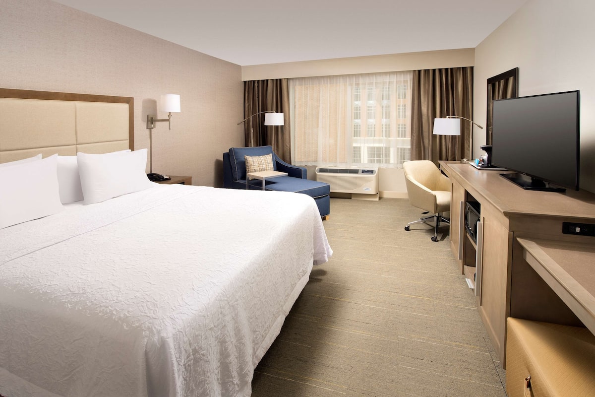 A hotel room with a large bed, a desk with a flat-screen TV, a chair, a desk lamp, and a window with curtains.