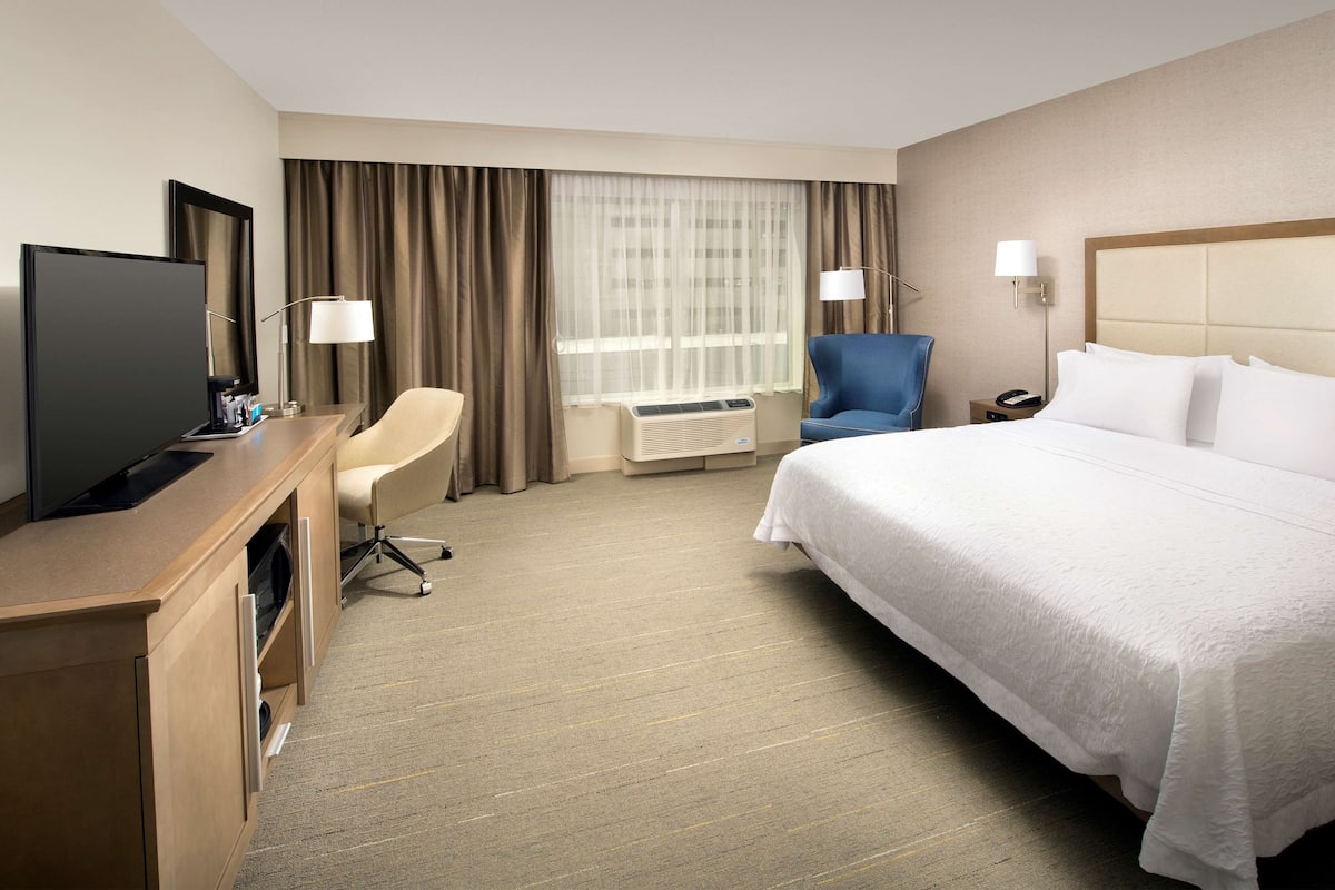 A hotel room with a bed, a desk with a television, a chair, and a window with curtains.